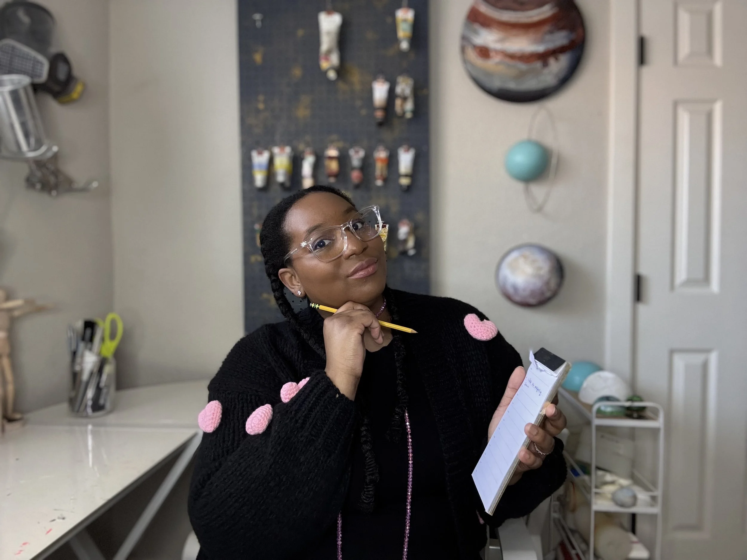 A woman with glasses and braids wearing a black sweater with pink pom-poms, holding a notepad and pen, standing in a room with art supplies and artwork on the wall behind her.