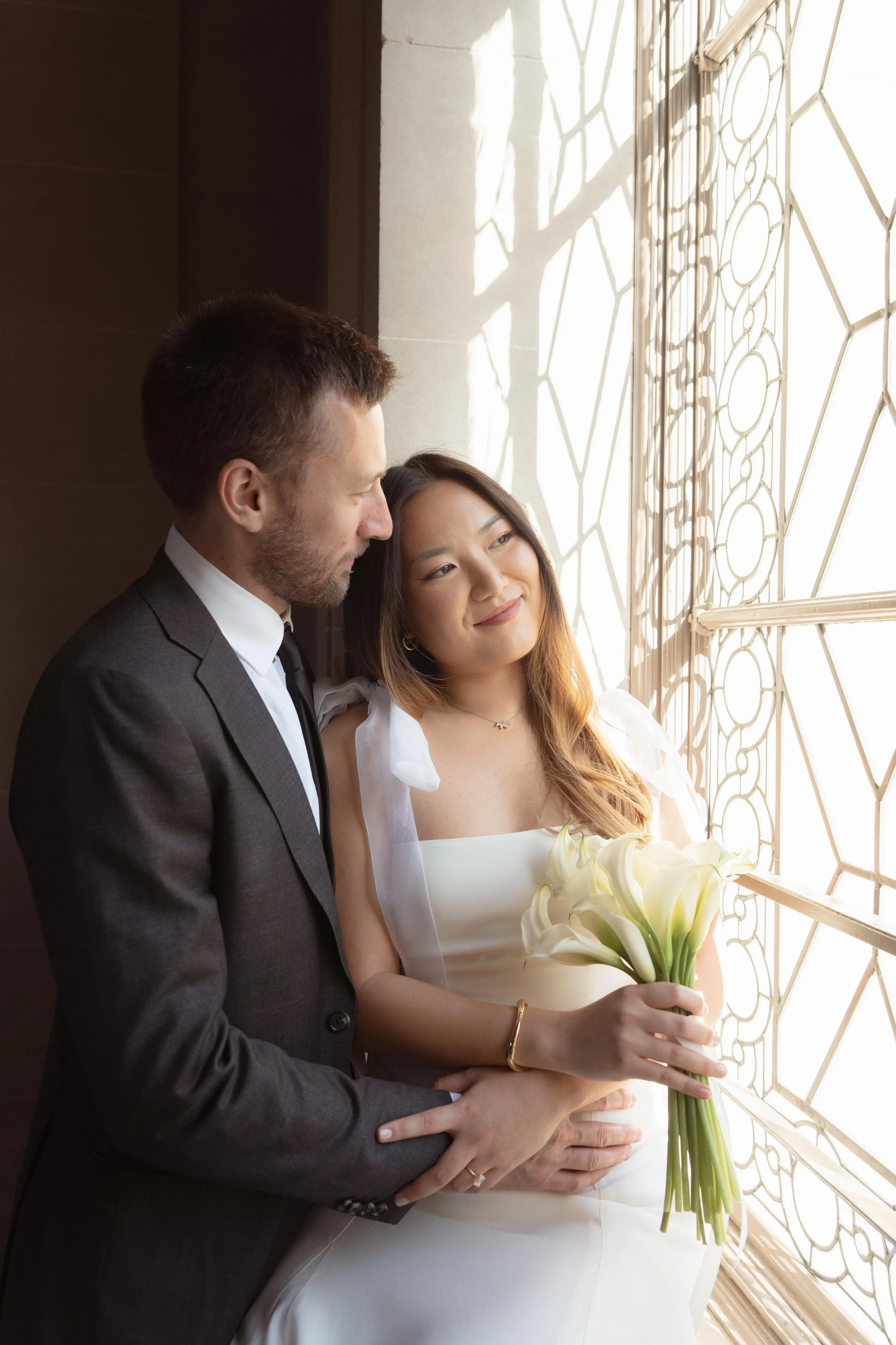 Couple at San Francisco City Hall courthouse wedding ceremony, holding white calla lily bouquet by a sunlit window