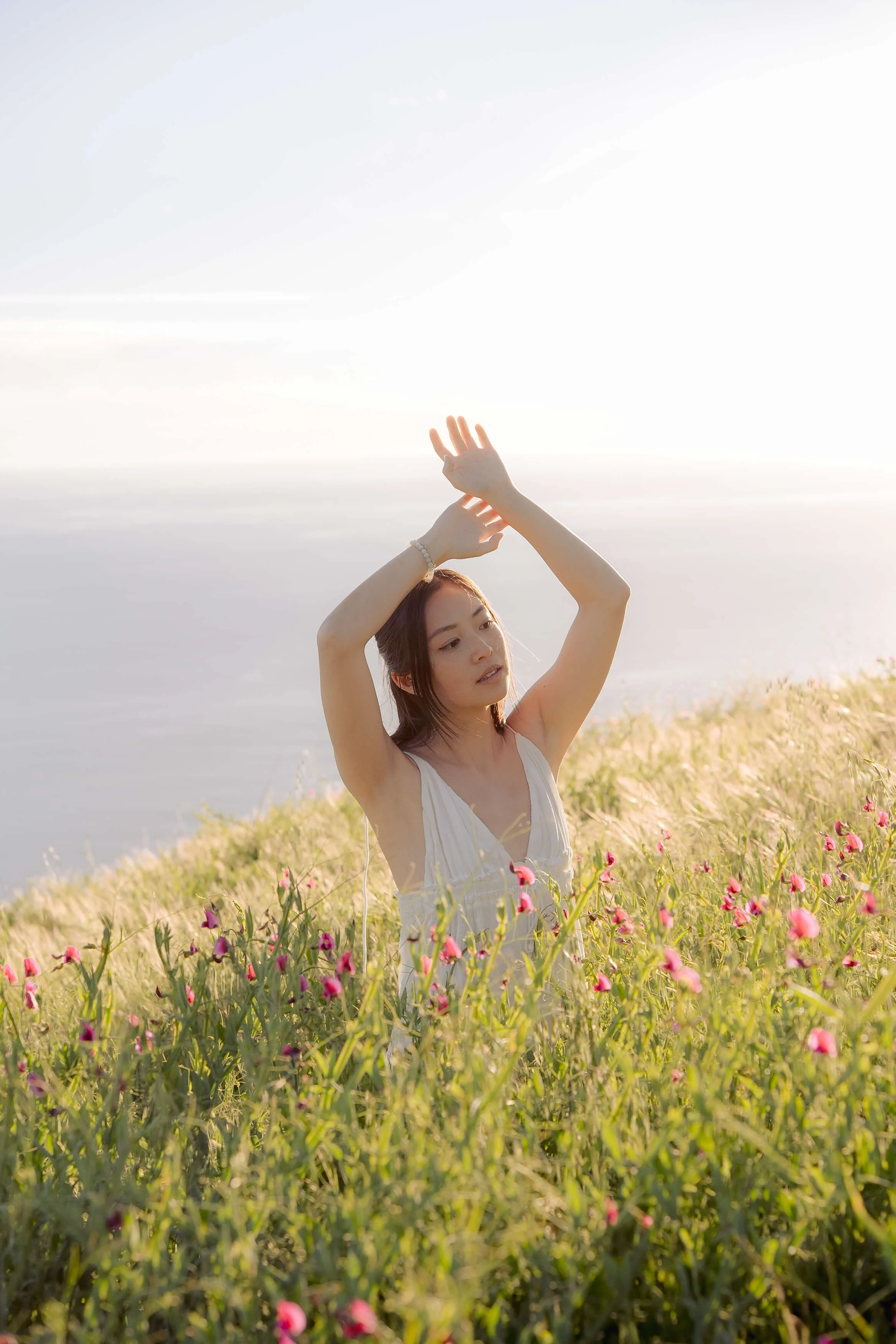 Editorial-style portrait of an Asian woman in a white dress standing in wildflowers by the ocean in Big Sur