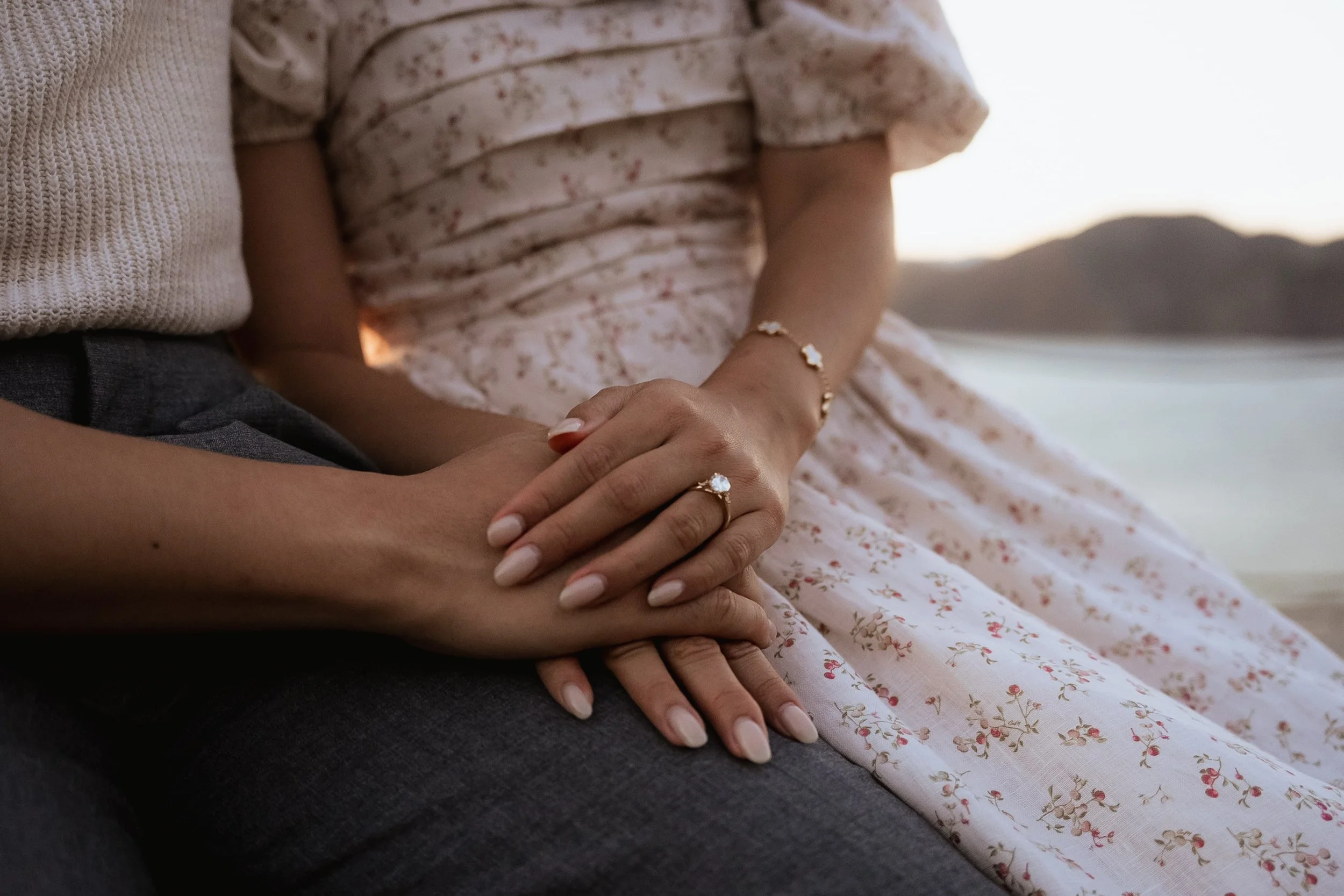 Sunset engagement photo of a couple holding hands by the water on the Batteries to Bluff trail in San Francisco, featuring a close-up of an engagement ring and the woman in a floral dress