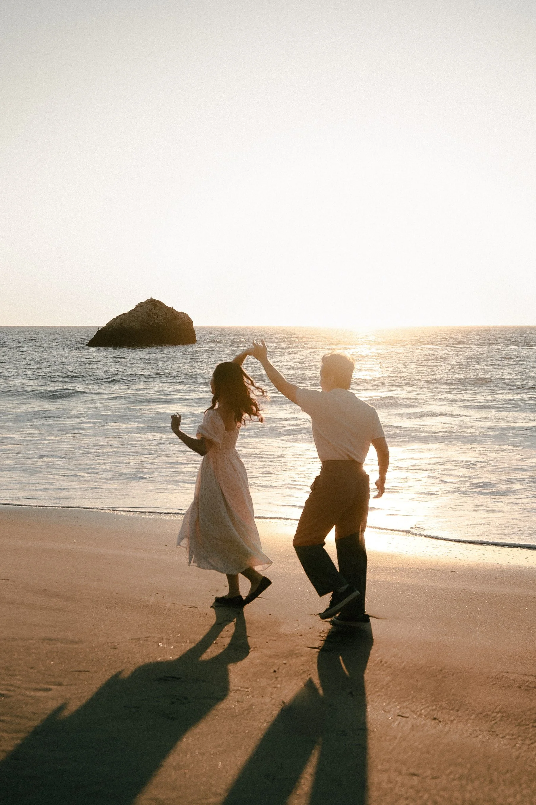 A couple dancing at Marshall's Beach in San Francisco during a sunset engagement photoshoot, with the ocean and large rocks in the background