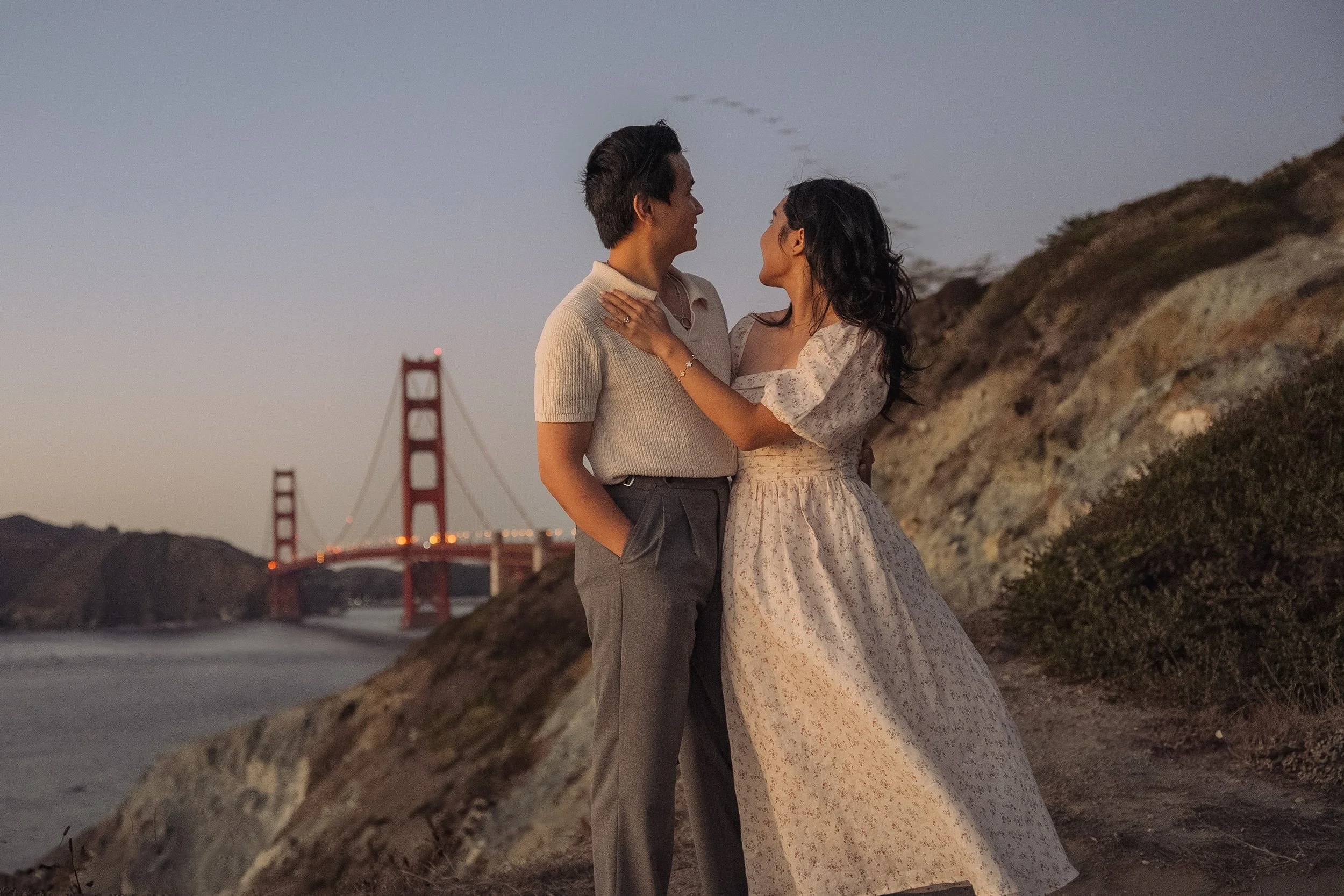 Sunset engagement photo of a couple at the Batteries to Bluff trail, standing on a hillside by the ocean at sunset, with the Golden Gate Bridge in the background
