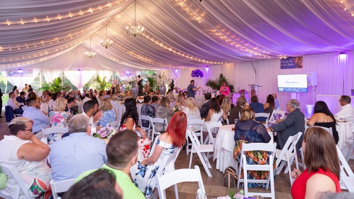 Indoor event with people seated at round tables under a tent with draped fabric and chandeliers, featuring a speaker on stage.