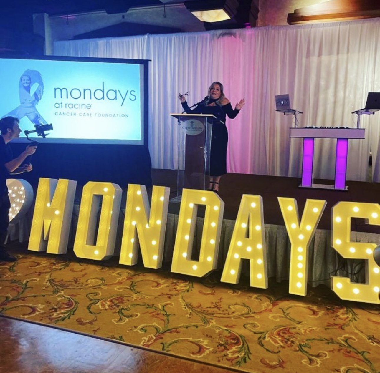 A woman speaking at a podium in a decorated event space, with a large display of the word 'MONDAYS' in illuminated letters. A banner shows 'Mondays at Racine Cancer Care Foundation'. Stage lighting and decor visible.