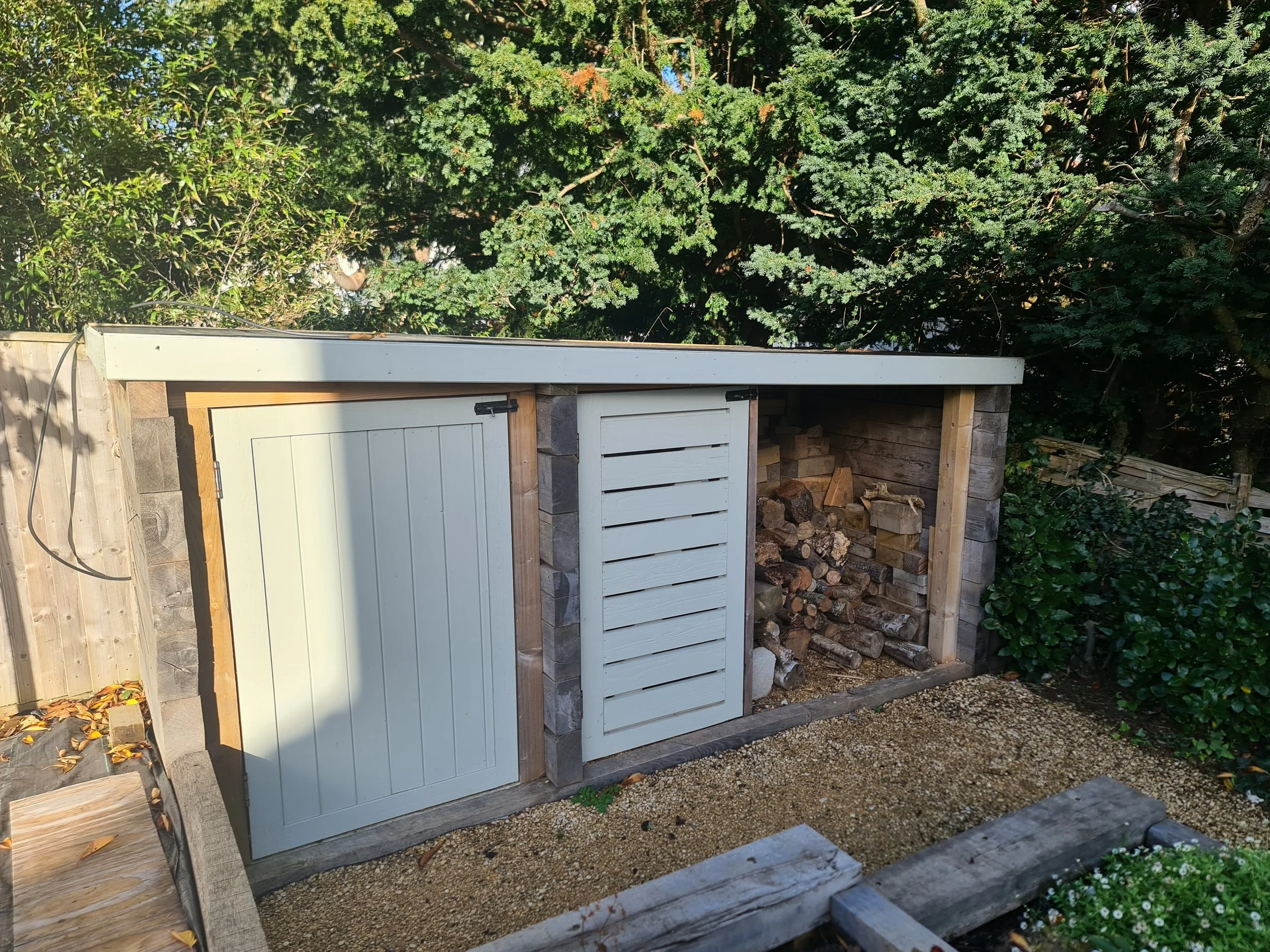 Wood storage shed with a white door and a ventilation panel, filled with stacked firewood, in a garden with trees and bushes in the background.