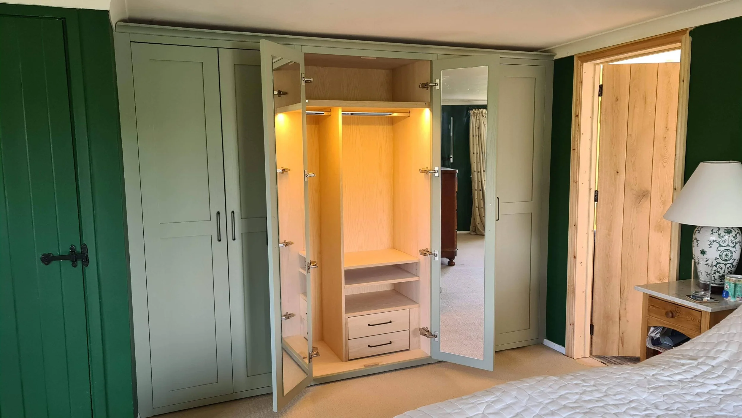 Open wardrobe with beige interior, small drawers, and hanging space, in a bedroom with green walls, wooden door, bedside table, and bed with white quilt.