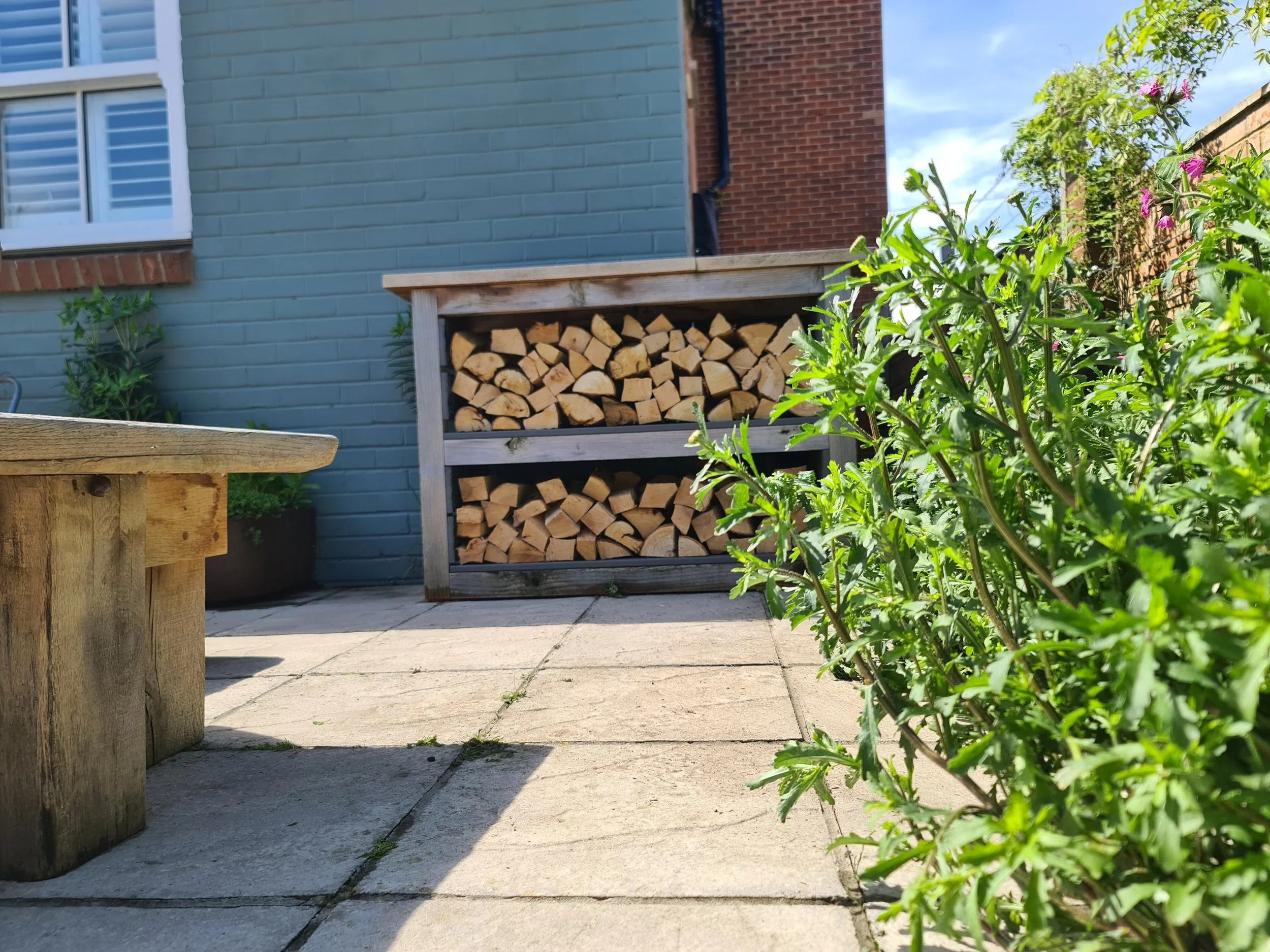 Backyard patio with a stack of chopped firewood in a wooden storage shelf, a small potted bush, green plants, and a house wall painted in blue and red bricks, under a partly cloudy sky.