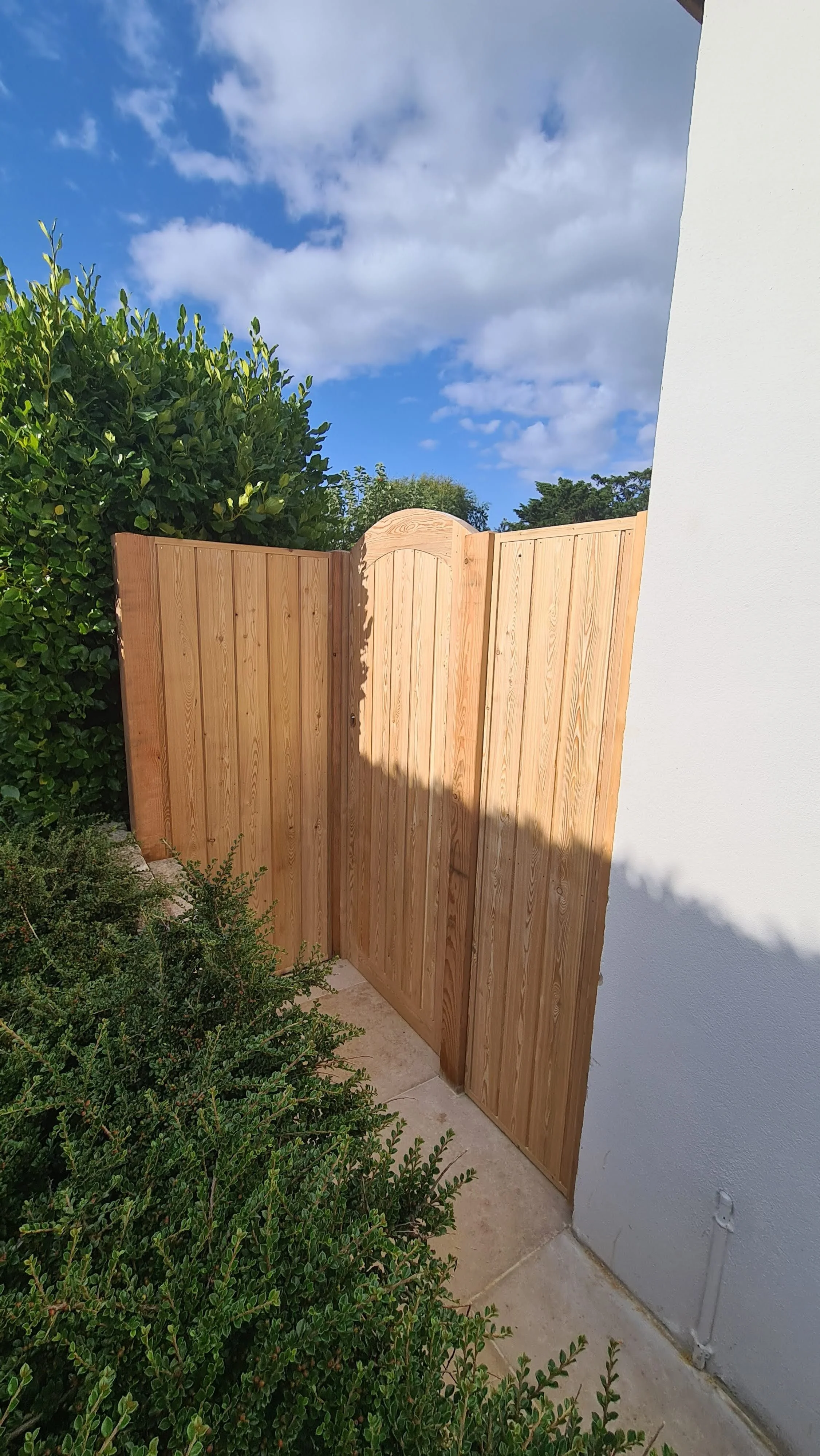 A newly installed wooden fence with a gate next to a white stucco wall, with green bushes and a blue sky with clouds in the background.