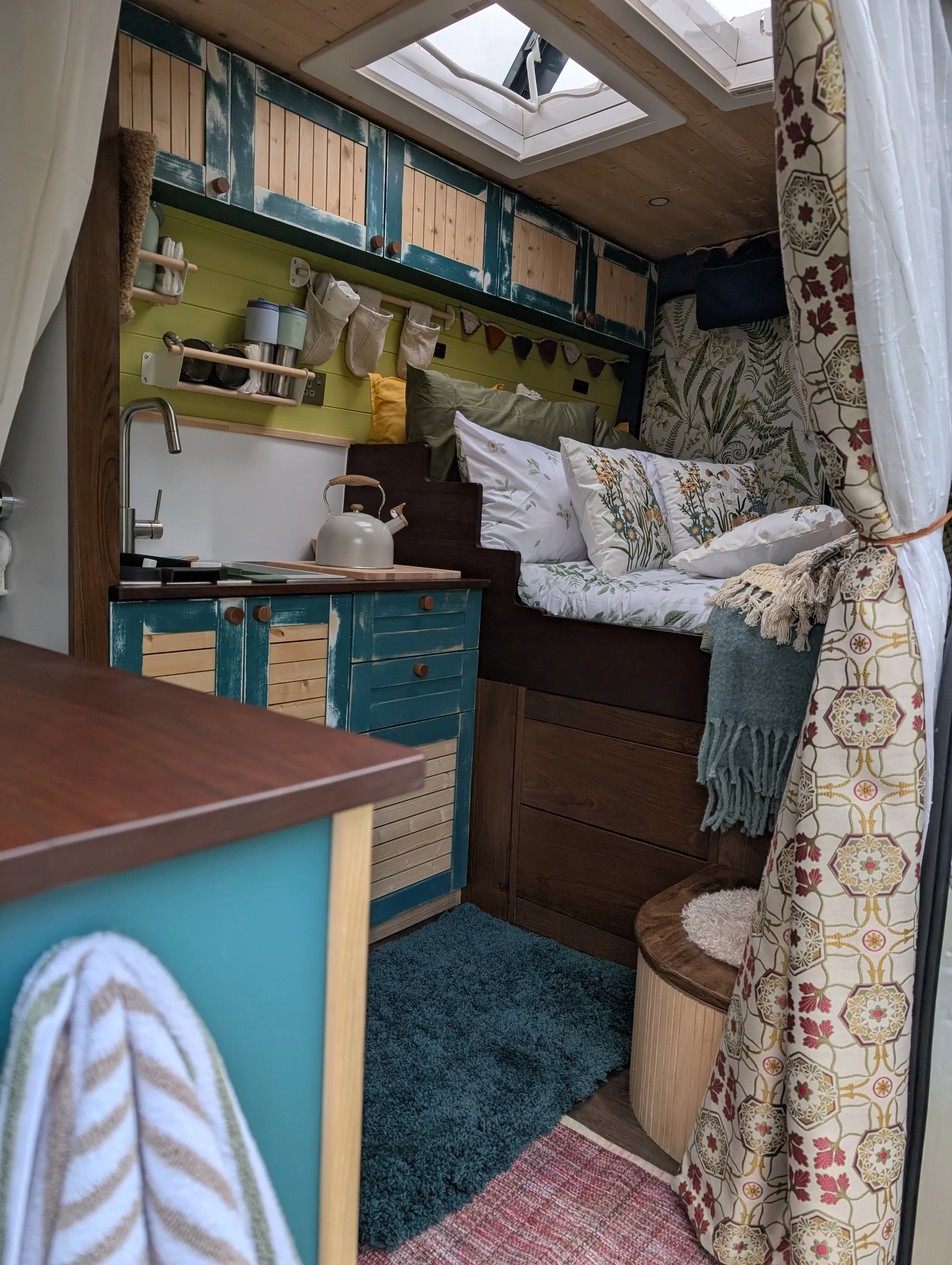 Cozy tiny home interior with a small kitchen area, bed with pillows, colorful decor, and a skylight overhead.