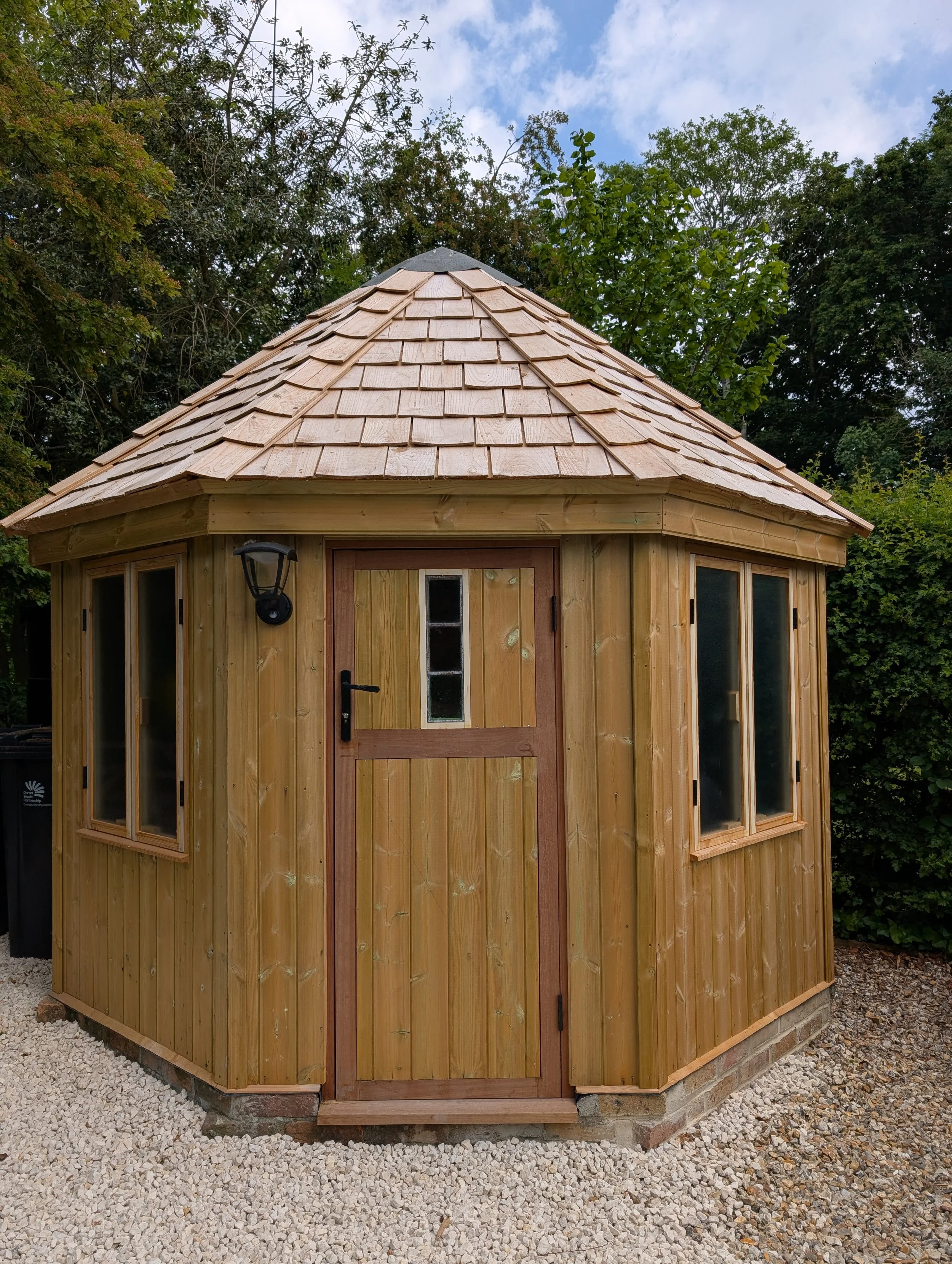 Small wooden garden shed with a shingled roof, featuring a door with a small window, two side windows, and an exterior lantern light, set on a gravel base.