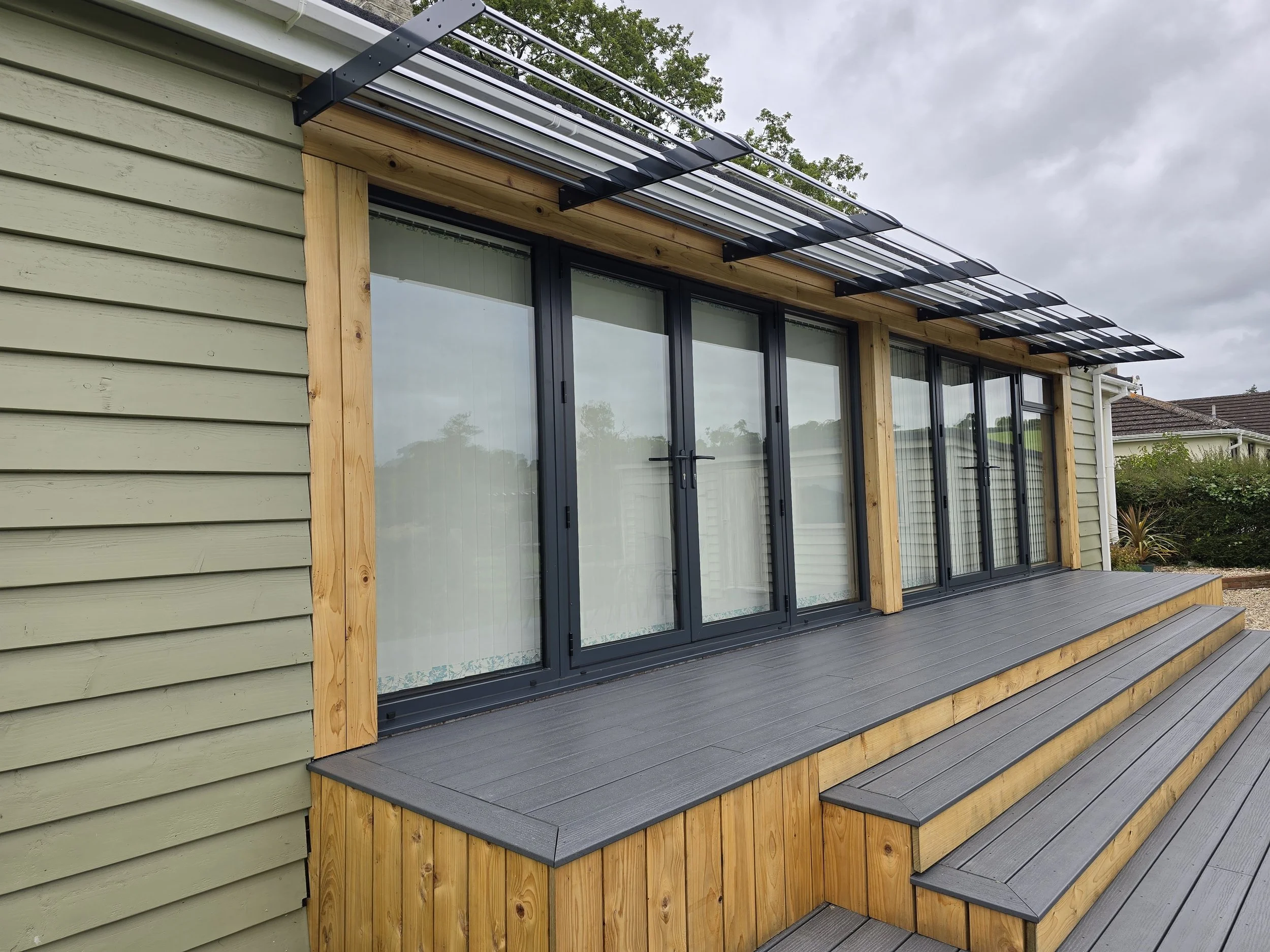A modern house exterior with large sliding glass doors, a composite and cedar deck with stairs, and a metal and glass awning over the door, surrounded by greenery and an overcast sky.