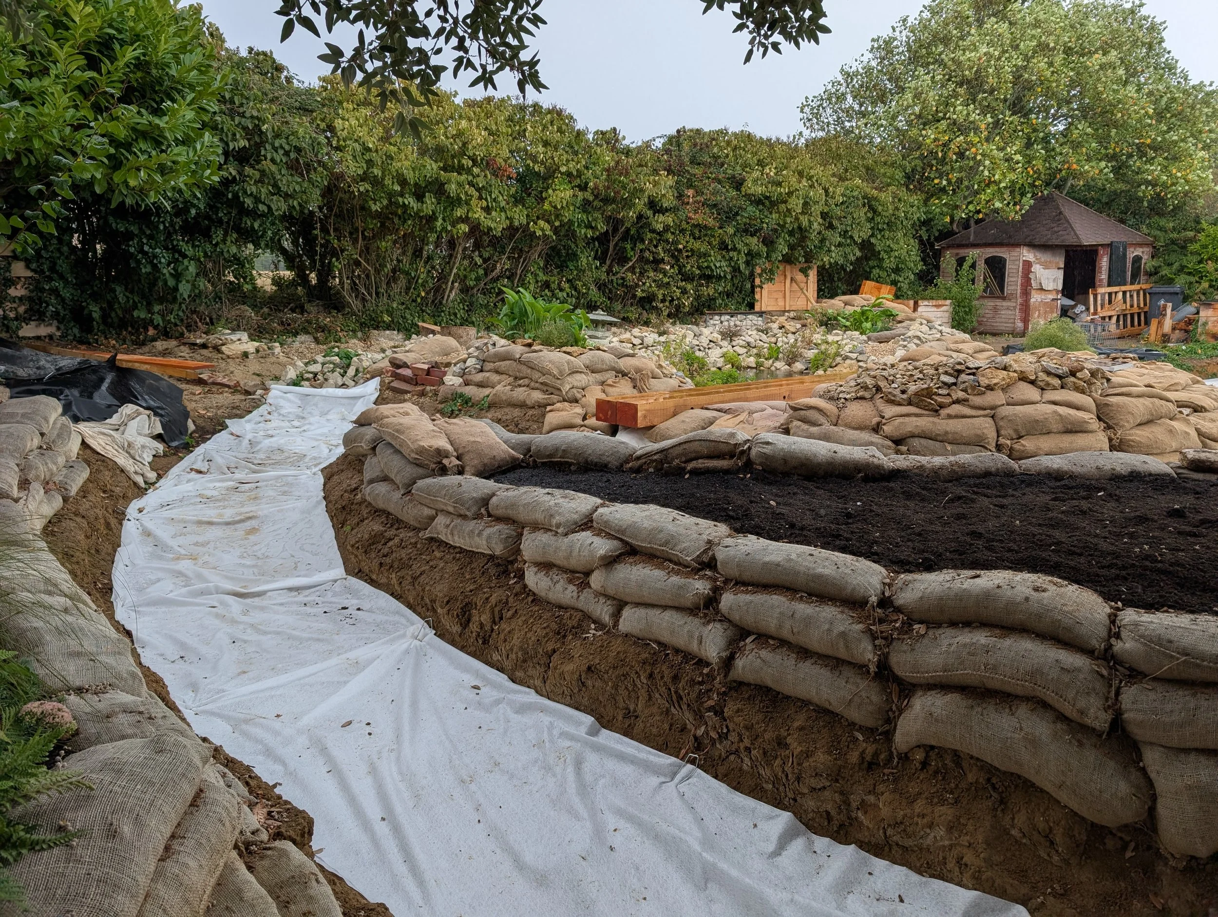 A garden or yard under construction, with dirt and soil, sandbags lining a trench, white fabric on the ground, additional sandbags, and a small wooden shed in the background surrounded by trees.
