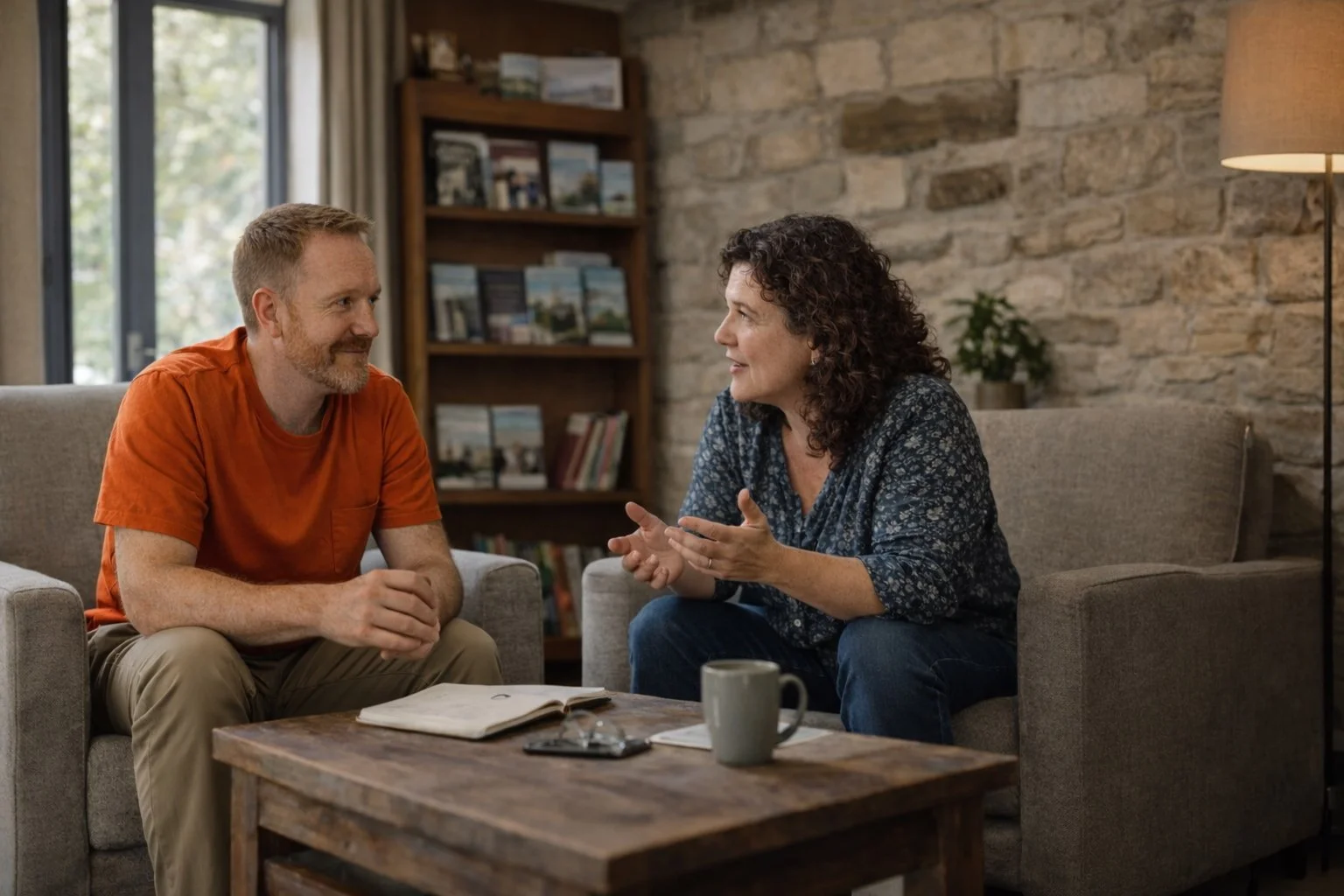 Two people sit facing each other in conversation inside a historic building, listening attentively and gesturing as they talk, with bookshelves in the background.