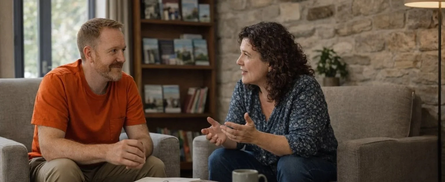Two people seated in armchairs, talking across a low table in a stone-walled room with bookshelves behind them.