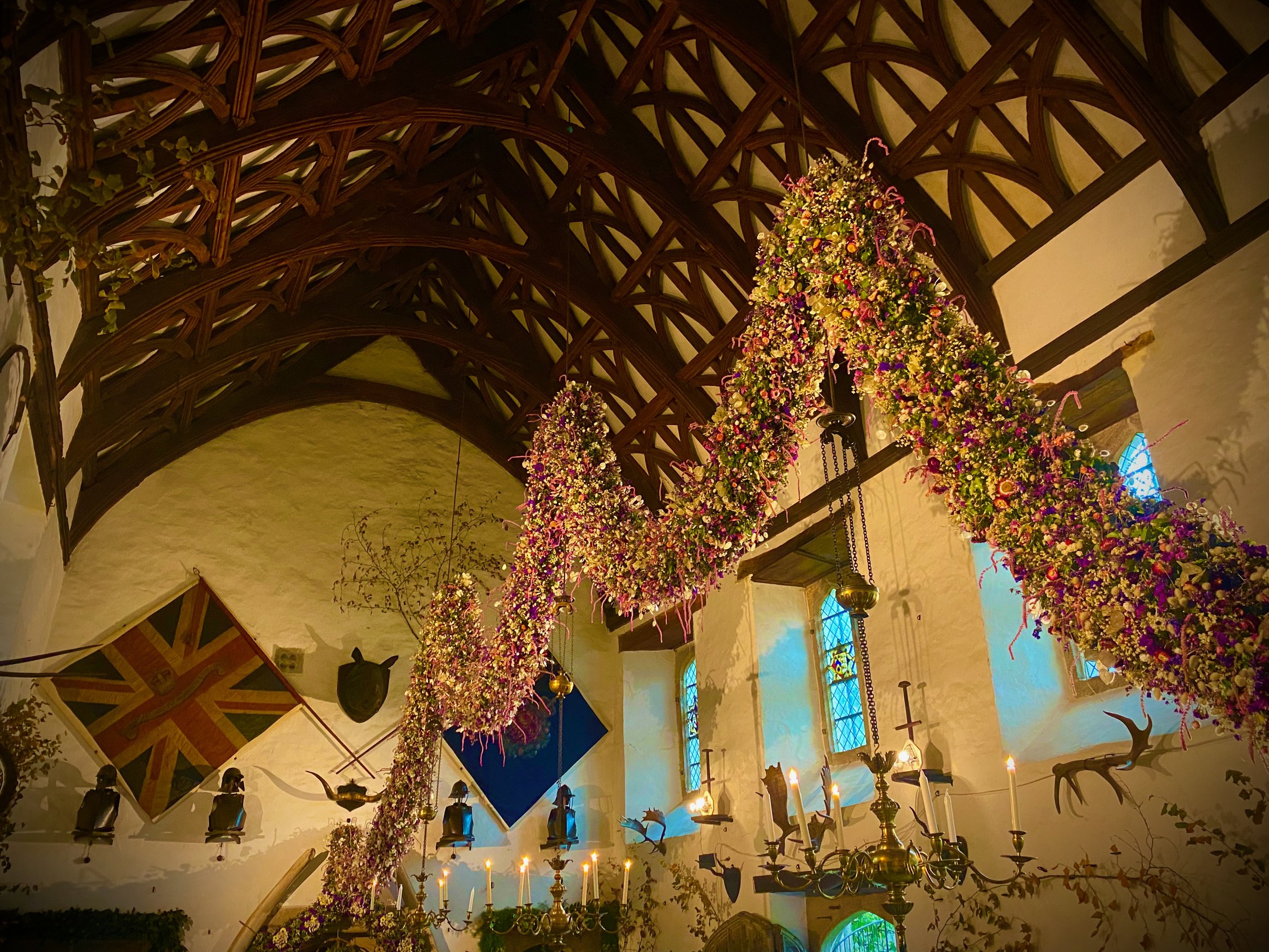 A vast floral Christmas garland hangs in sweeping curves beneath a timber-vaulted ceiling in the Great Hall at Cotehele, with chandeliers, candles, heraldic shields and stained-glass windows visible below.