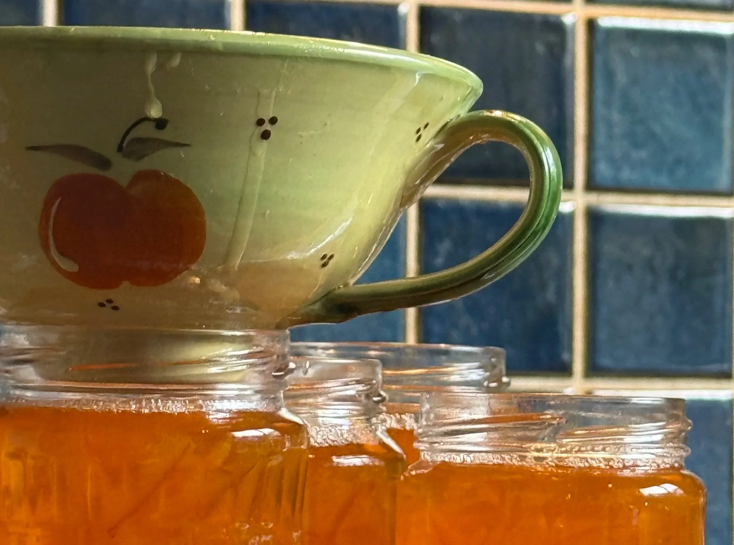 A ceramic funnel rests on the rim of glass jars as bright orange marmalade is poured into them in a kitchen with blue tiled walls