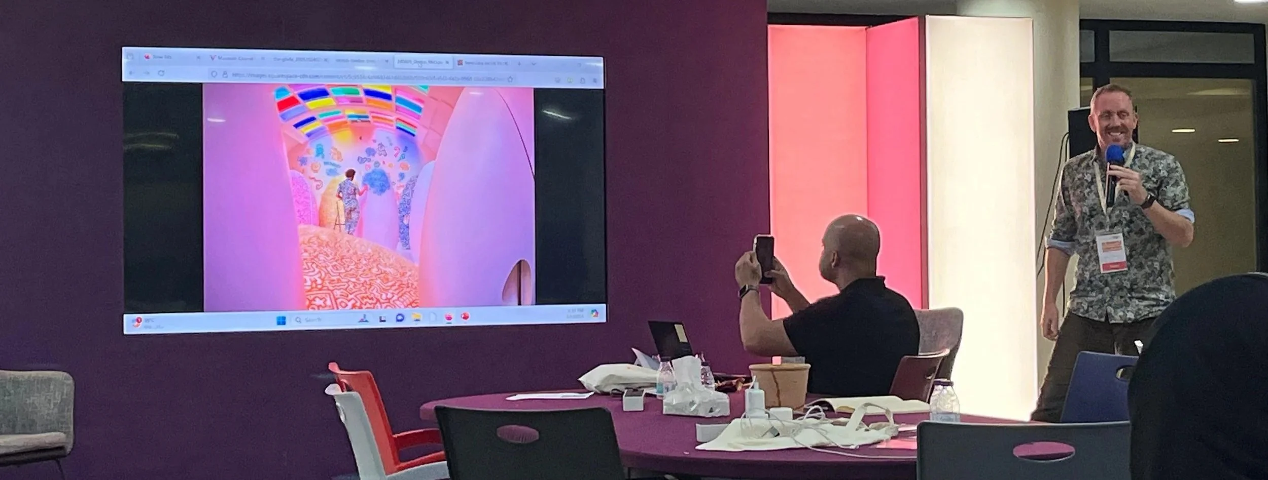 A person speaking into a microphone during a workshop, with a presentation screen visible behind them and seated attendees in the room.