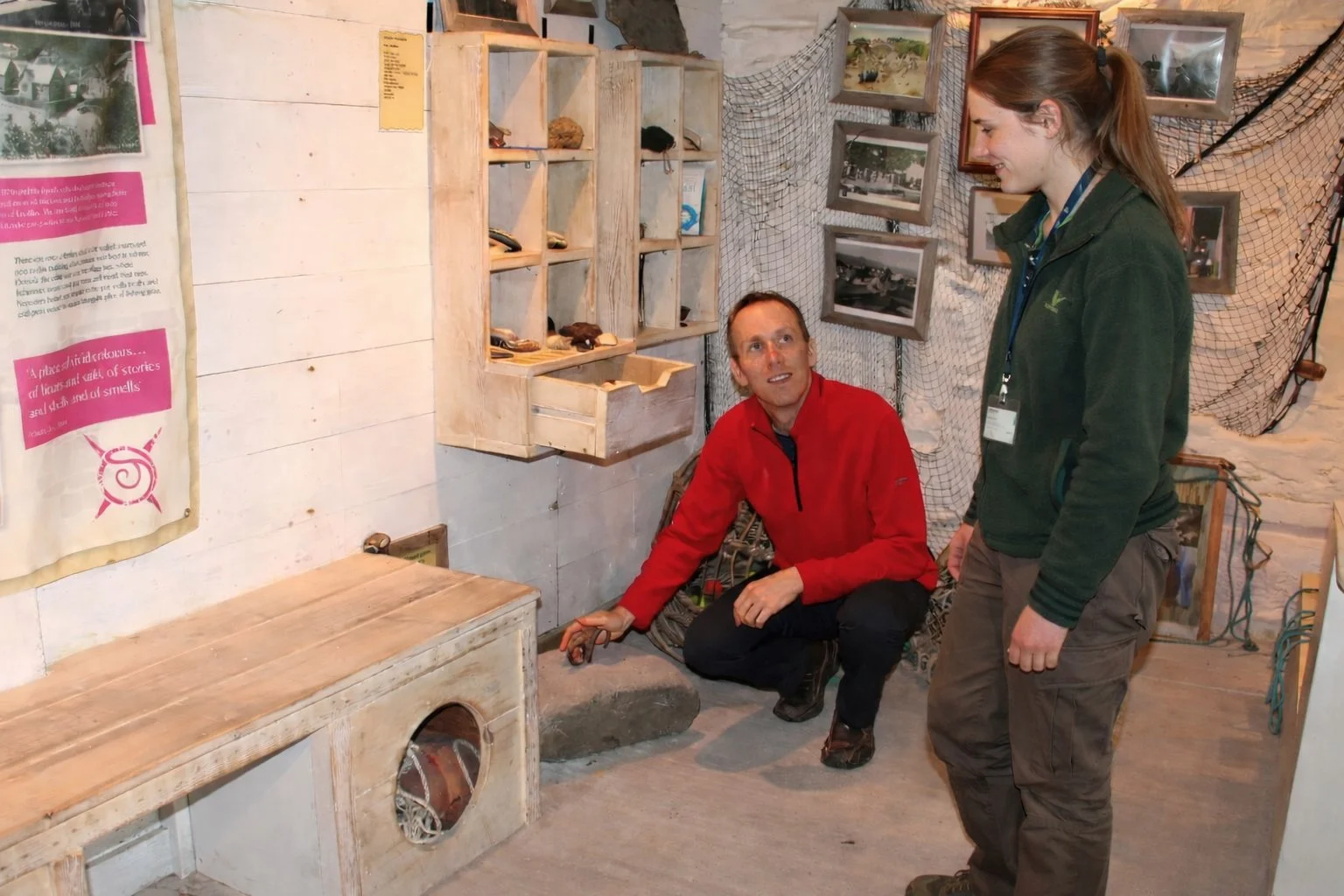 A member of staff and a colleague stand and crouch inside a small heritage display room, discussing an object set into a low wooden structure, with historic photographs and artefacts on the walls.