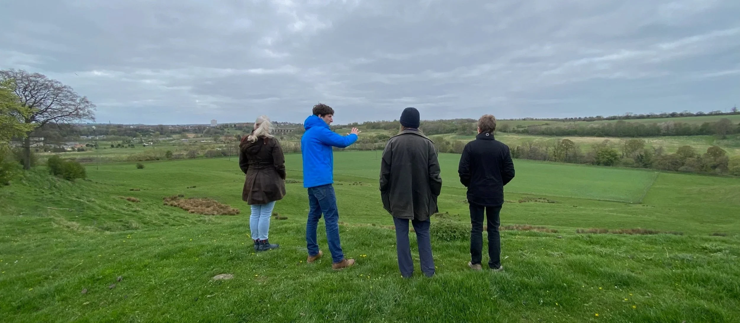 Four people on a grassy slope overlooking rolling farmland, one gesturing across the landscape as others look on.