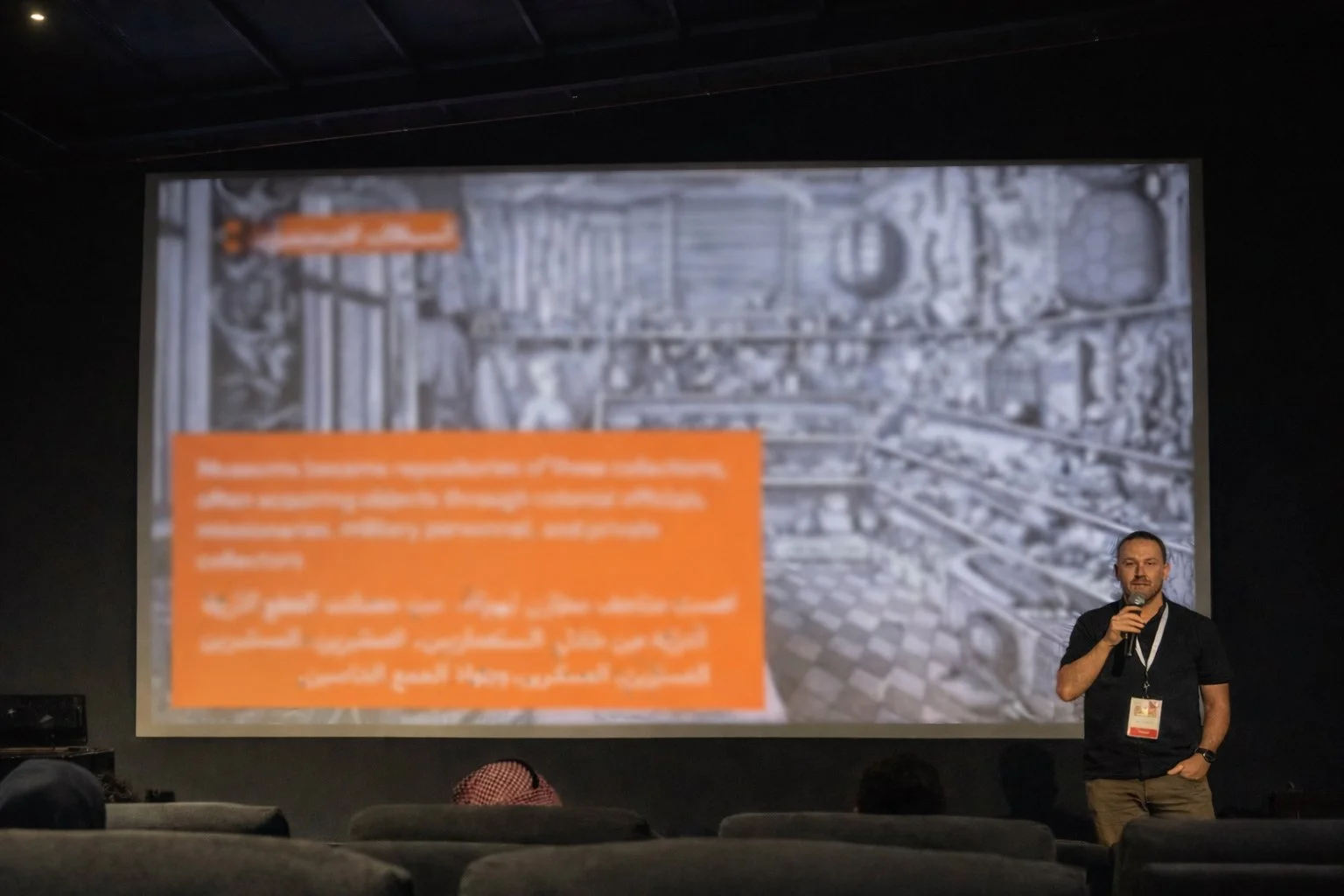 A speaker standing in front of a large projected slide during a presentation in a darkened auditorium.