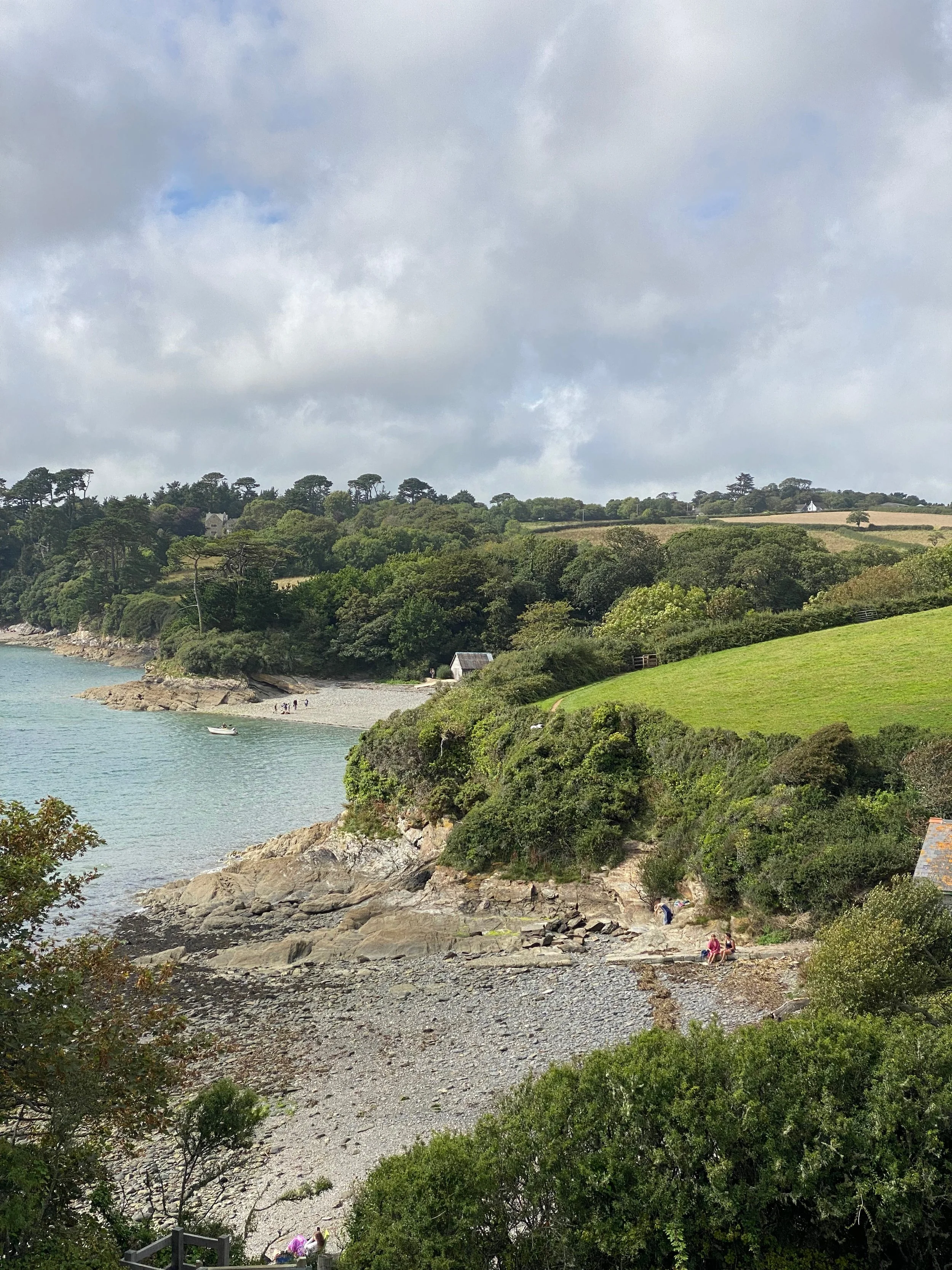 Rocky Cornish cove with calm sea, small boat offshore and scattered people on the beach, backed by wooded cliffs, patchwork fields and soft grey cloud above.
