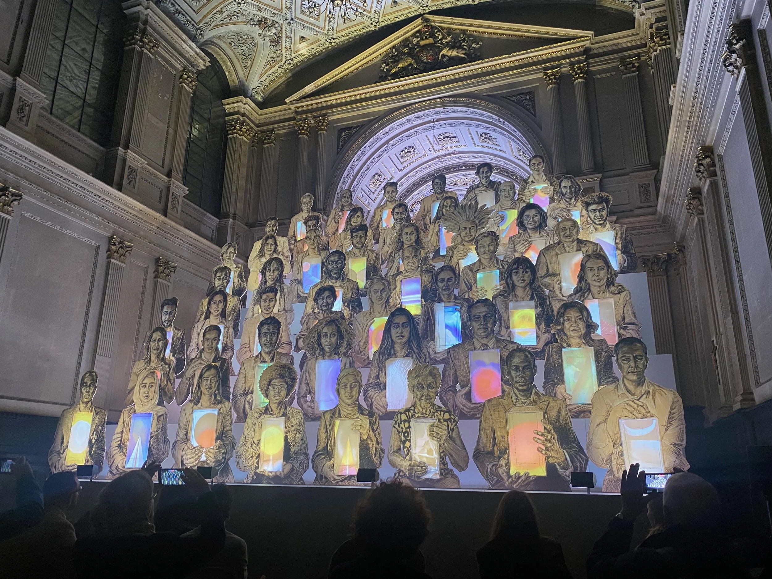 Two dimensional drawings of a number of different people are lined up in a rows one above the other. Each person holds a lit up colourful placard with shapes but no words on them.