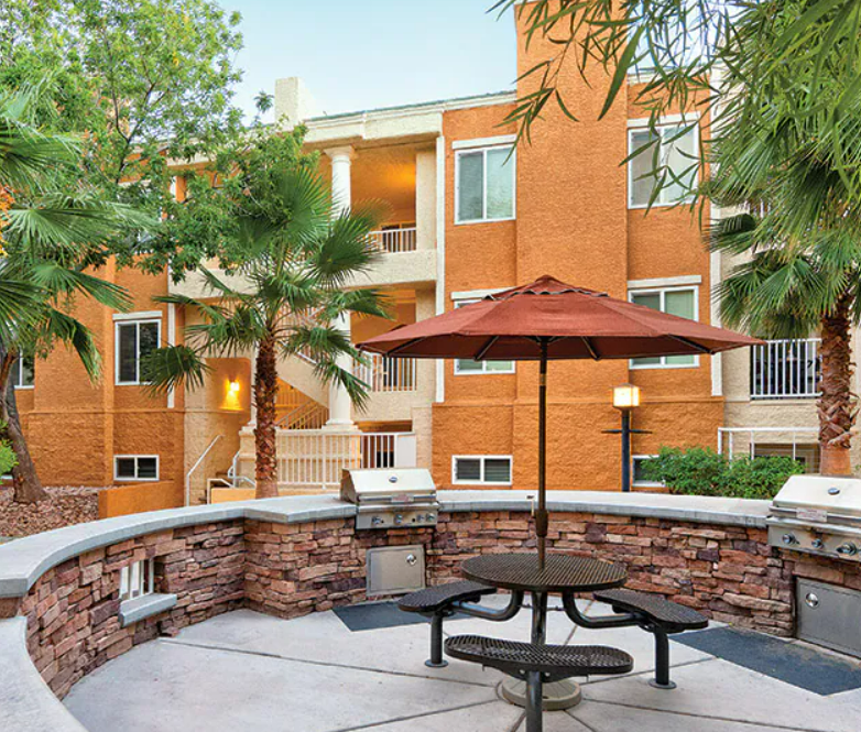 Outdoor patio area with a round table, attached umbrella, and two grills, in front of an orange residential building with balconies and lush greenery.