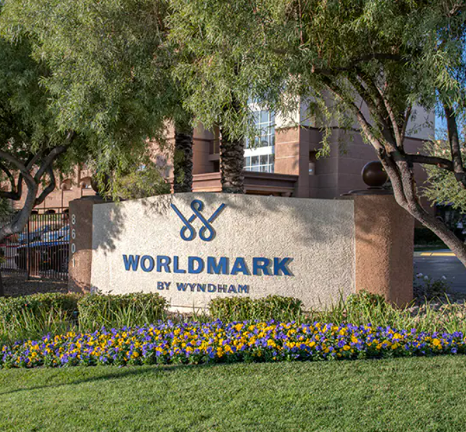 Sign for WorldMark by Wyndham with scissors logo, surrounded by flowers and trees.