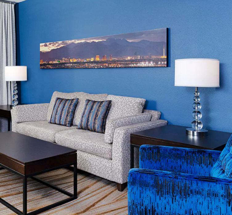 Living room with a white patterned sofa, blue armchair, dark wood side tables, white table lamps, a landscape wall art, blue walls, and a striped carpet.