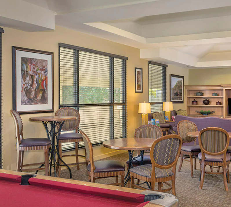 A cozy lounge area with round tables and upholstered chairs, large windows with blinds, framed artwork on the walls, and a bookshelf in the background.