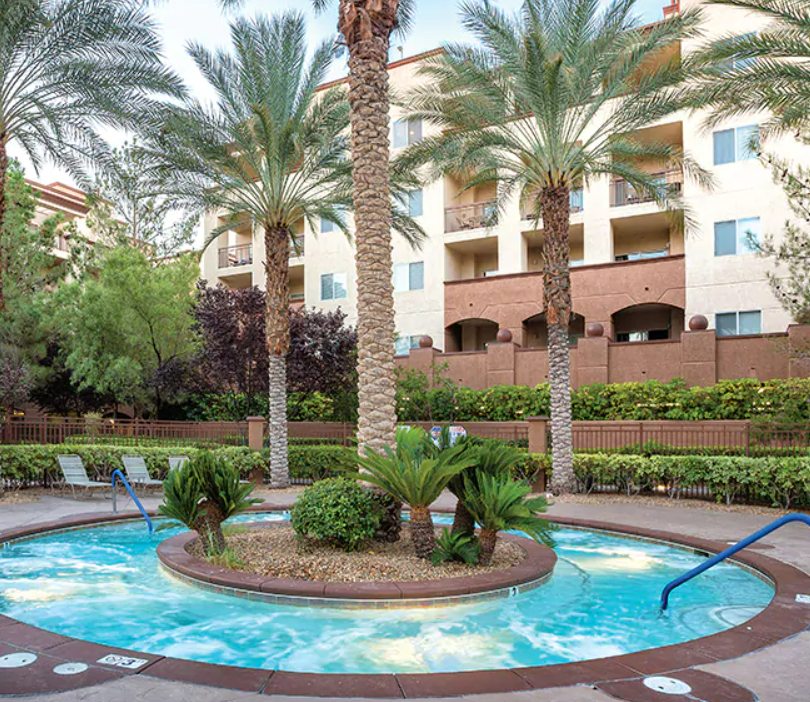 Swimming pool area with palm trees, lounge chairs, and a multi-story residential building in the background.