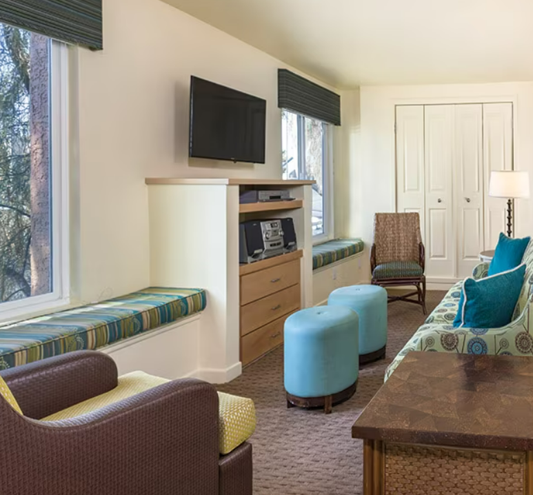 Living room with a wall-mounted TV, large windows with blinds, a patterned sofa with blue accent pillows, two blue ottomans, a brown armchair, and a small table with a lamp.