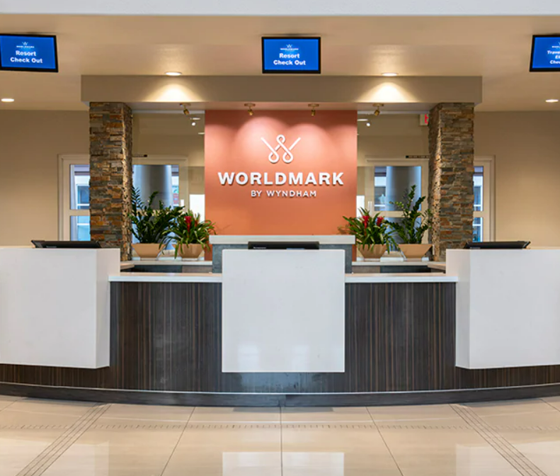 Hotel reception desk at Worldmark by Wyndham, featuring a sign with the logo, potted plants, and digital check-in screens overhead.
