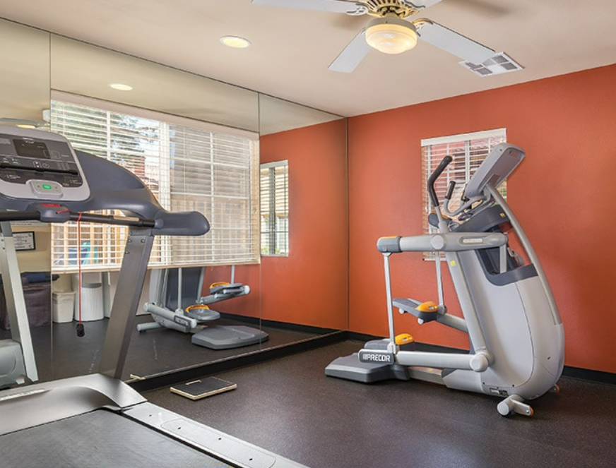 Home gym with treadmill and elliptical machine, large mirror on wall, ceiling fan, and window with blinds.
