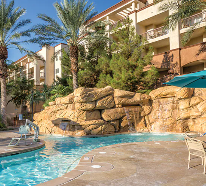 Swimming pool with rock waterfall feature, surrounded by palm trees, lounge chairs, and an apartment building in the background on a sunny day.