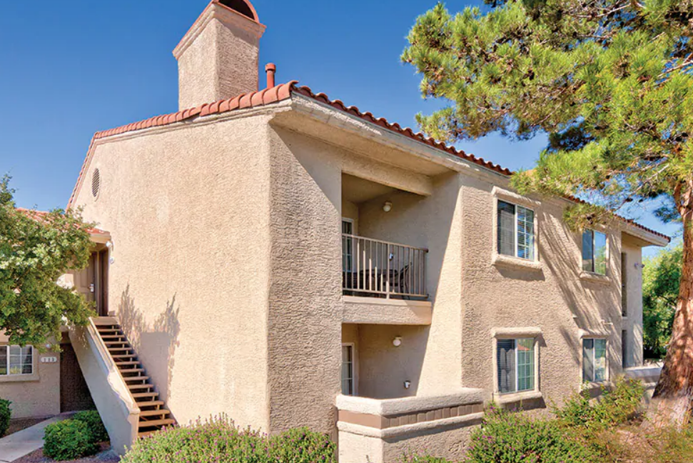 Two-story beige apartment building with a red-tiled roof and an outdoor staircase, surrounded by trees and bushes, under a clear blue sky.