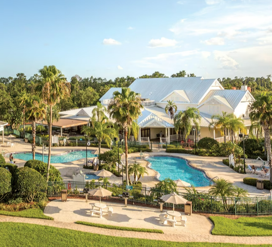 Swimming pool area with palm trees, lounge chairs, umbrella shades, and a large white clubhouse building surrounded by greenery under a partly cloudy sky.