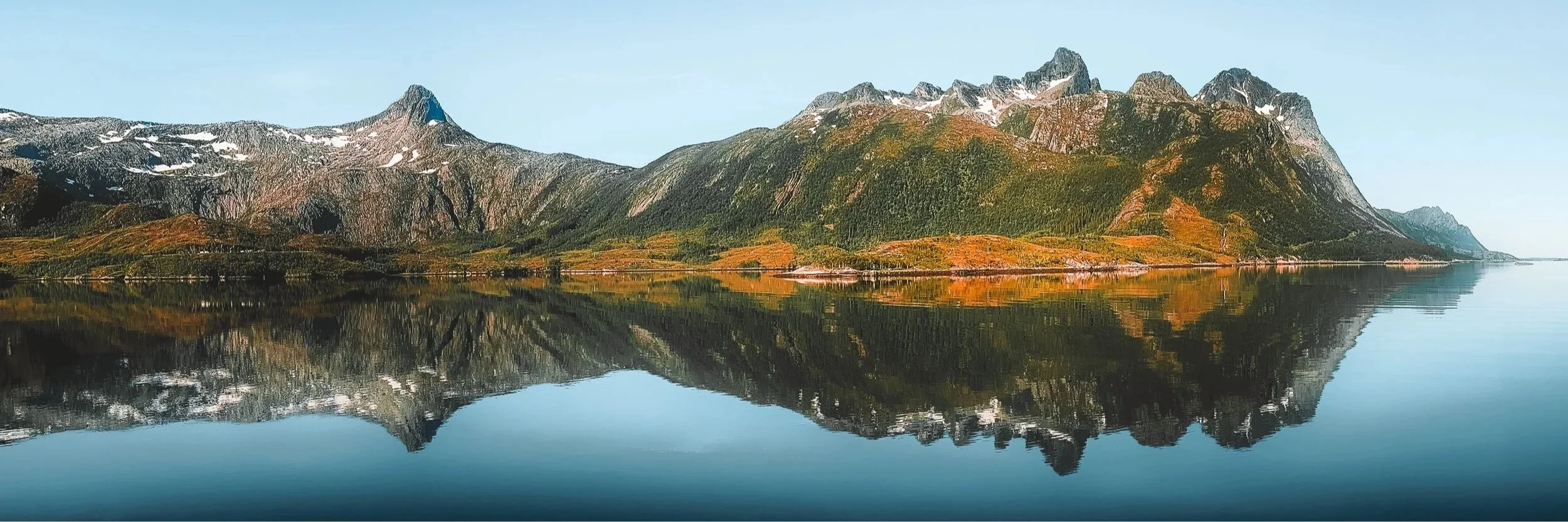 Panoramic view of mountains reflected in calm water in Vestland, Norway, with autumn colours and clear sky.