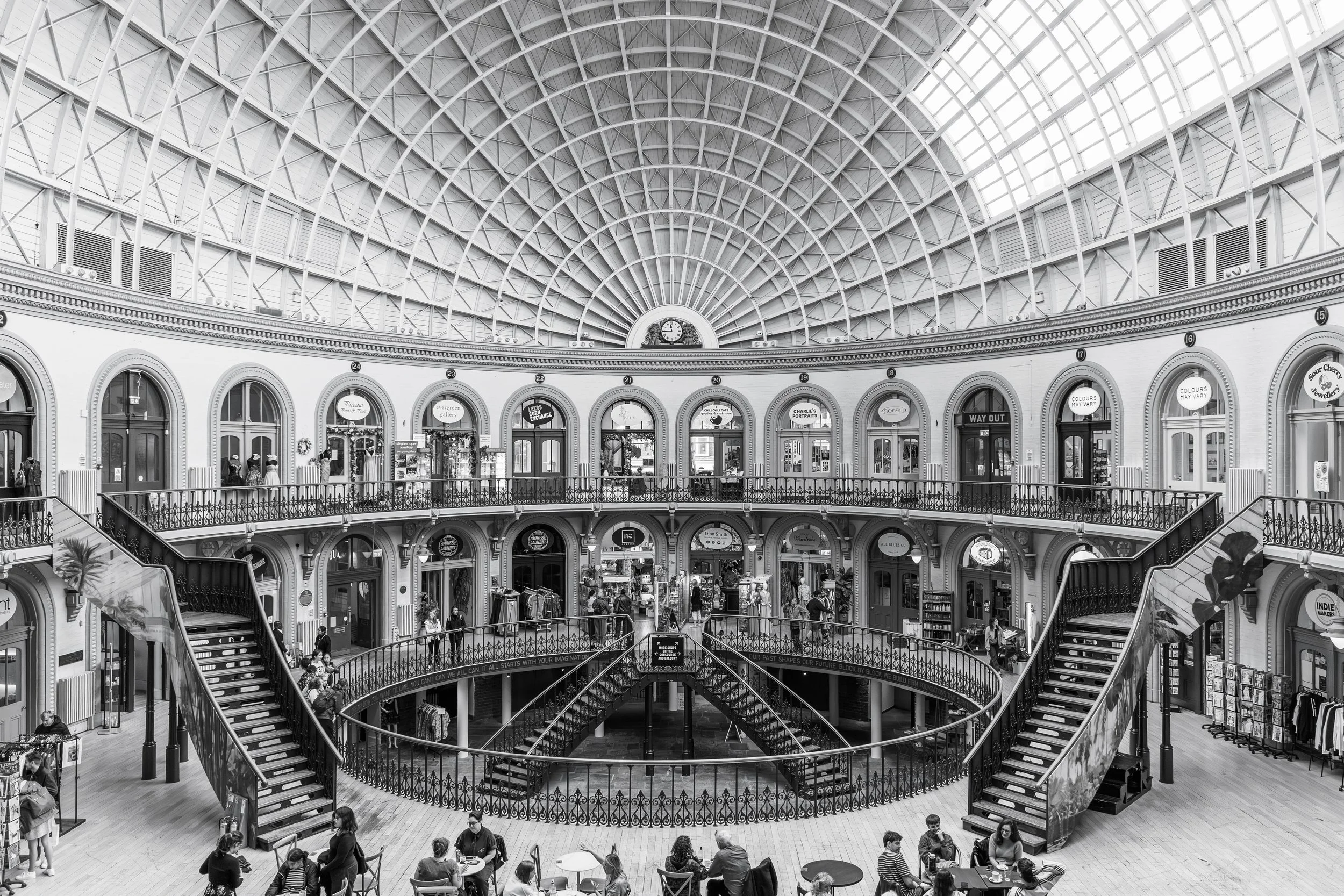 Black and white photograph of a grand indoor hall with glass roof, balconies, staircases and architectural detail.