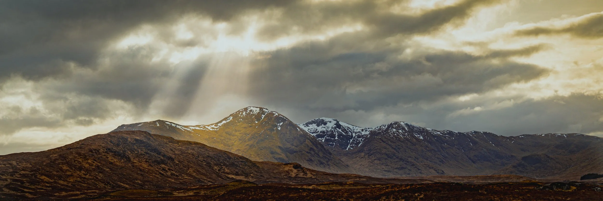 Panoramic view of sunlight breaking through clouds over Rannoch Moor and mountains in Scotland.