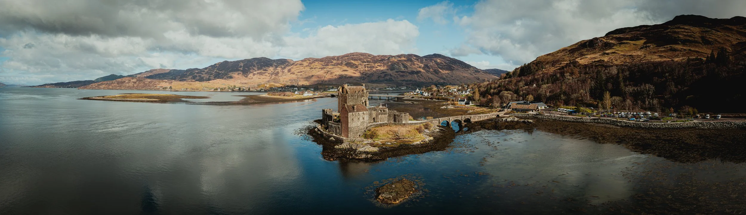 Aerial panoramic view of Eilean Donan Castle in Scotland with water, bridge, hills and cloudy sky.