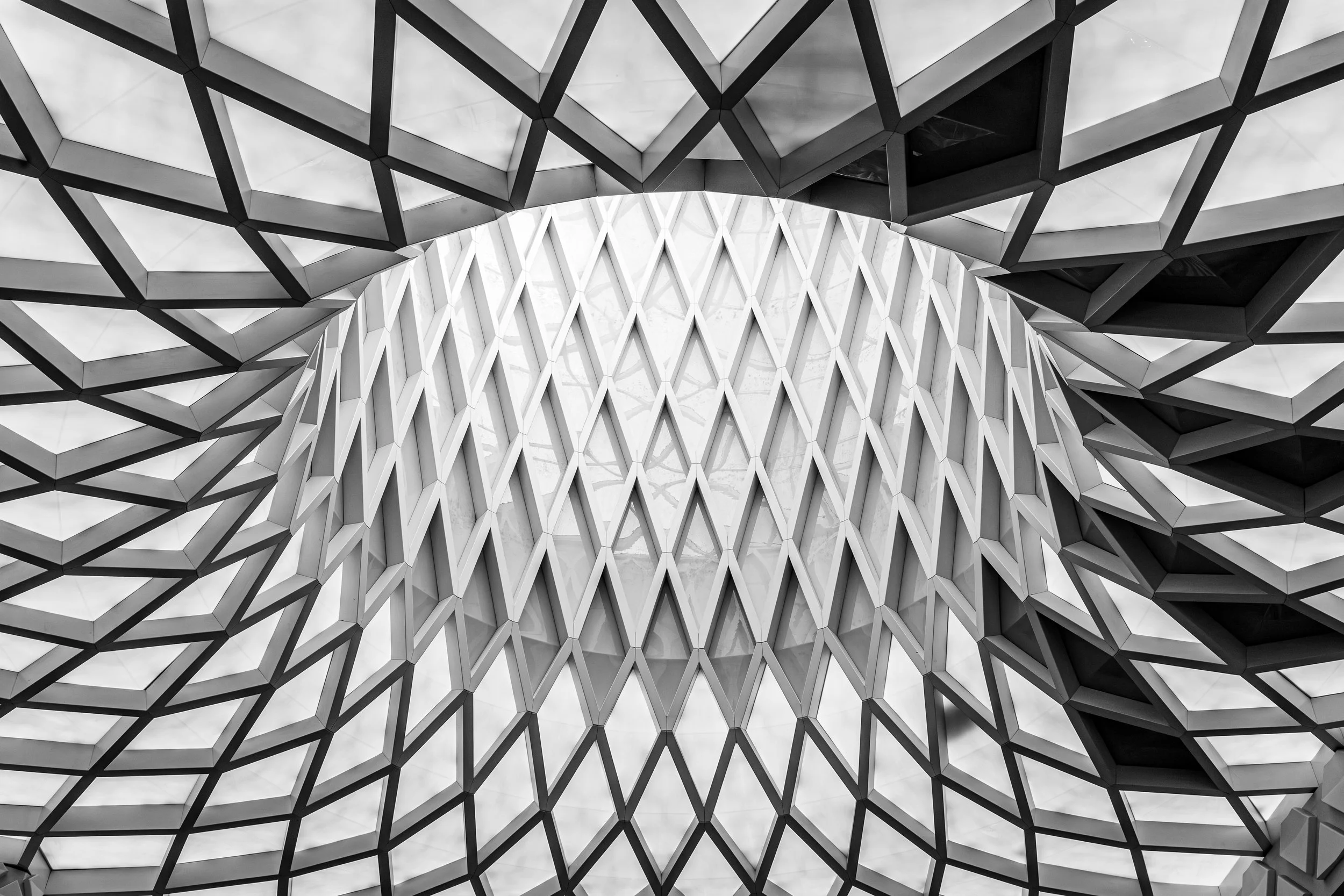 Black and white architectural photograph looking up into a modern glass and steel structure with repeating diamond patterns and a central funnel shape.