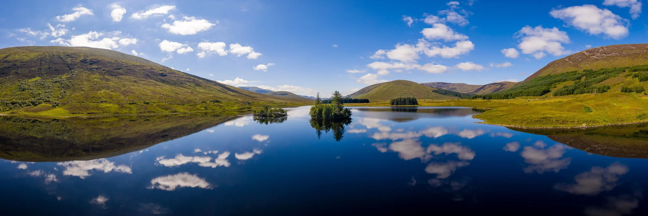 Panoramic view of Loch Scavon in Scotland with still water reflecting green hills, clouds and blue sky.