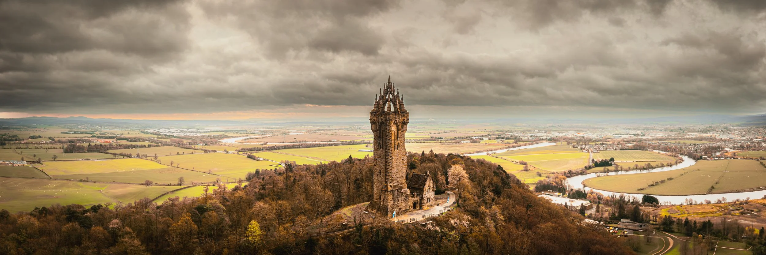 Aerial panoramic view of the Wallace Monument in Stirling, Scotland, with fields, hills and cloudy sky.