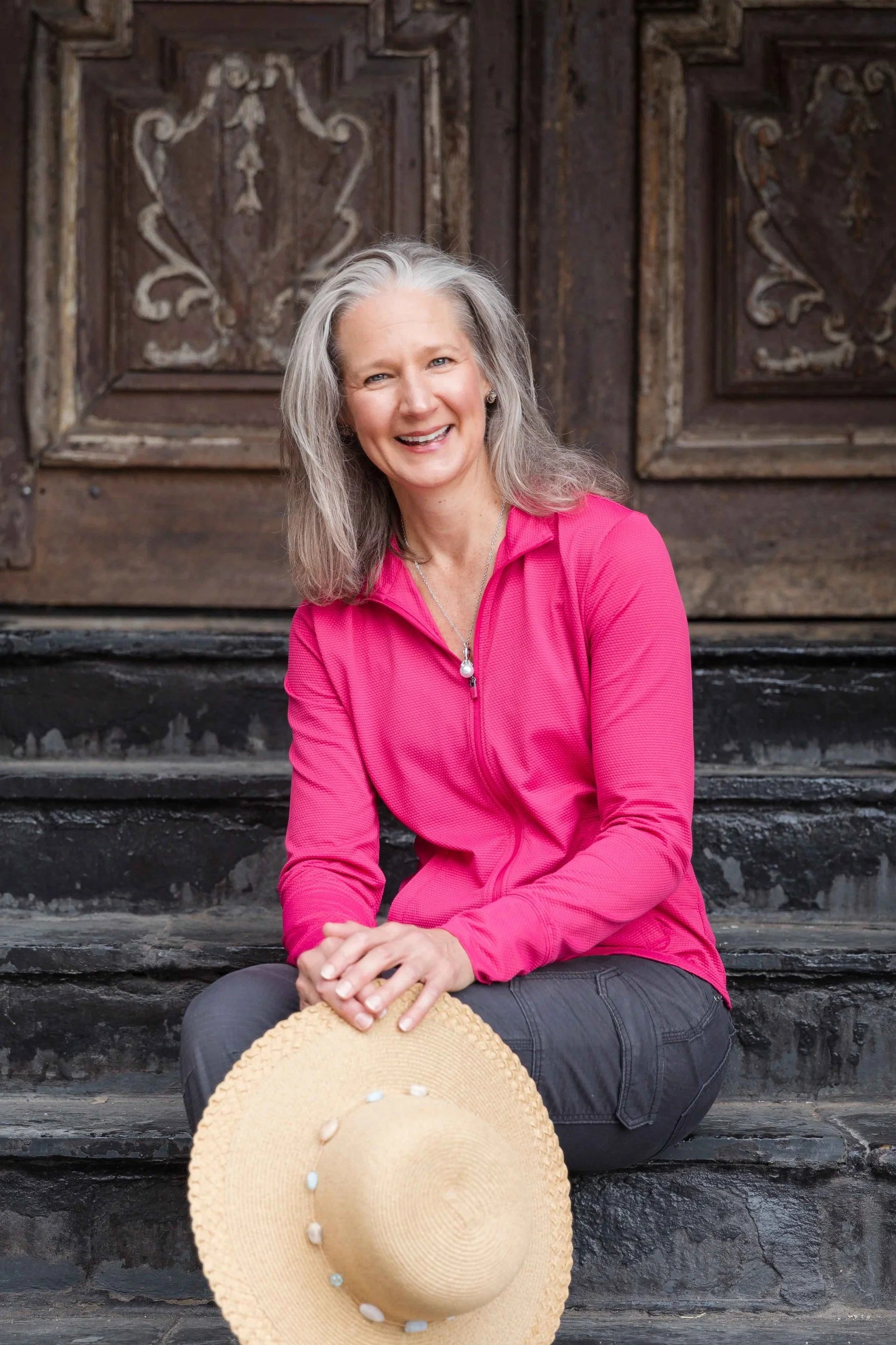 A smiling woman with gray hair sitting on black stone steps in front of a wooden door with decorative carvings, wearing a bright pink jacket, gray pants, and holding a straw hat with white decorations.