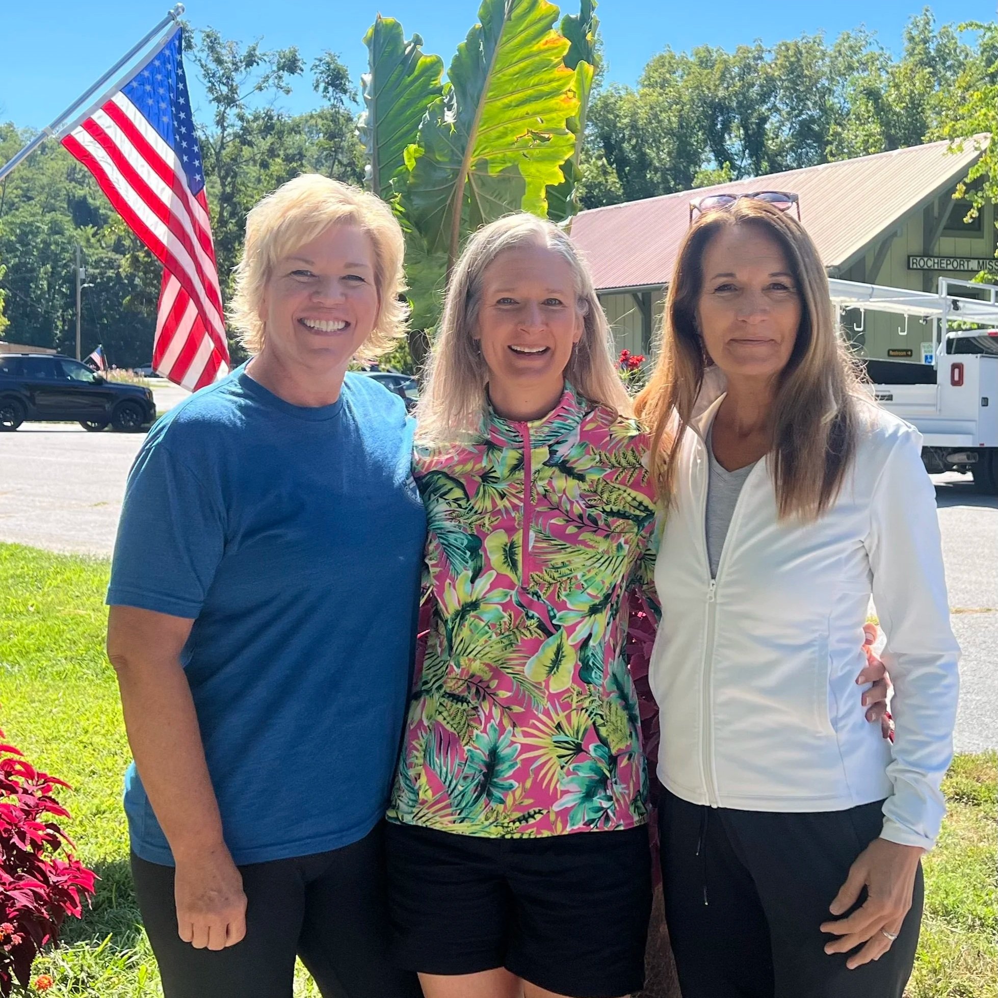 Three women standing outdoors in front of a large green leaf, an American flag, and a building with a roof. They are smiling and posing for the photo on a sunny day.
