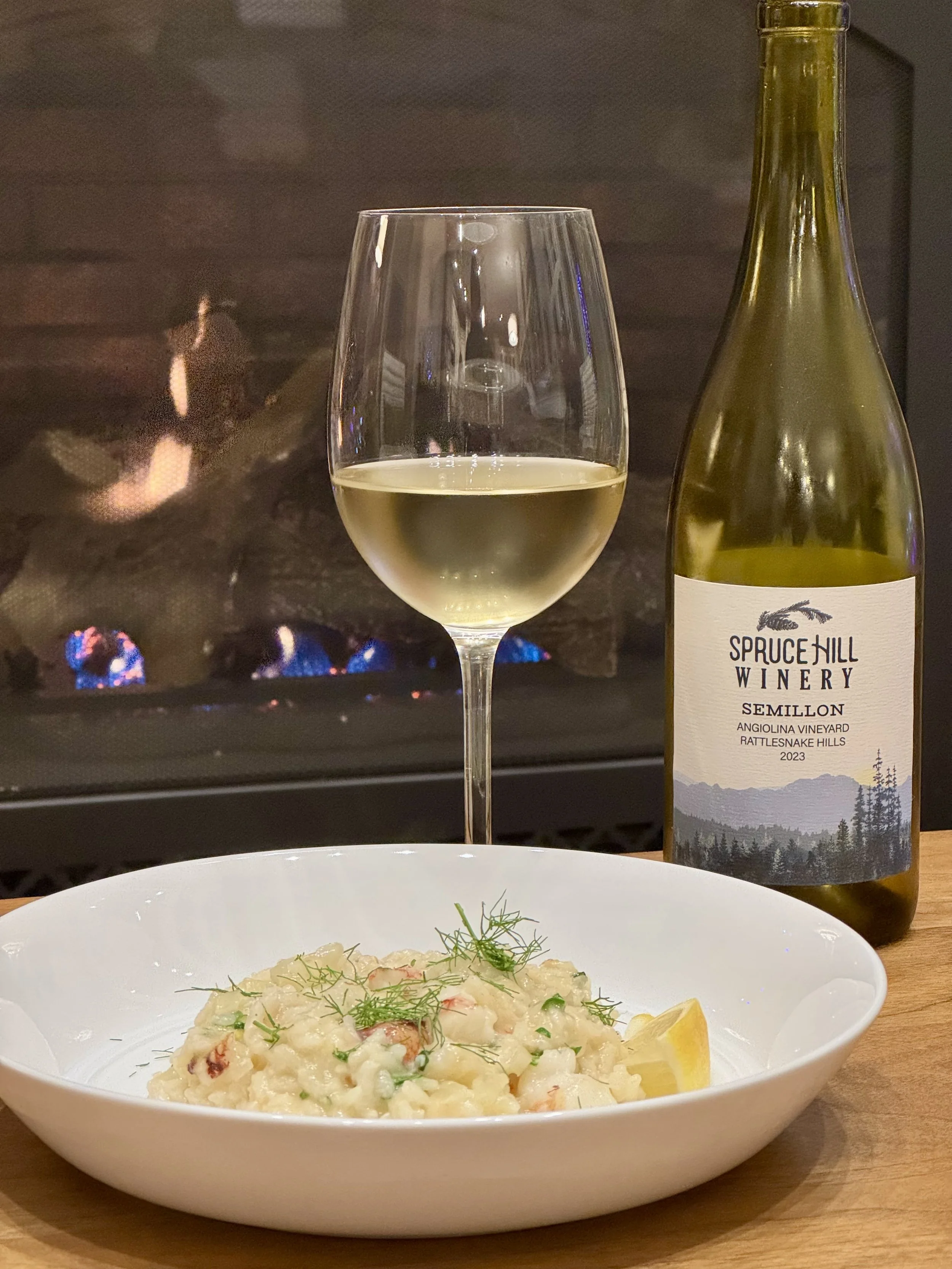A plate of risotto garnished with herbs, a glass of white wine, and a bottle labeled Spruce Hill Winery Semillon, placed in front of a warm fireplace.