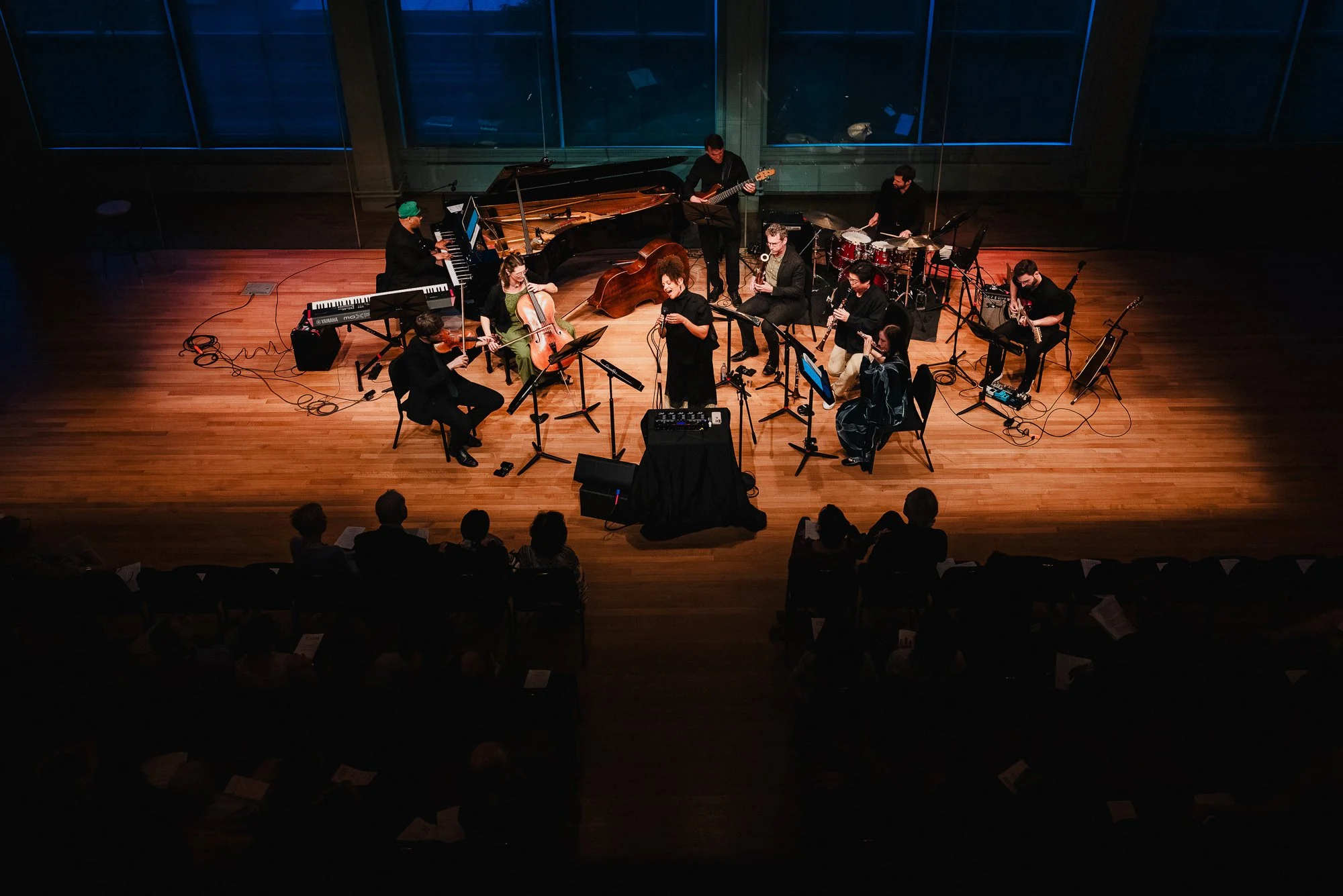 Overhead view of the full ensemble performing in Weill Music Room, audience visible on both sides, warm center light on the stage, windows behind the performers.