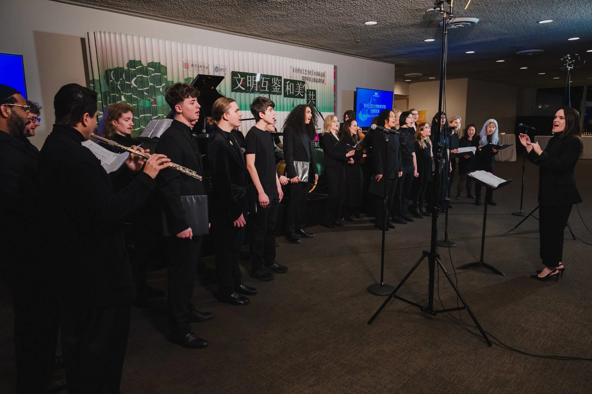 Teachers College Choir in all black performs at United Nations headquarters, conductor at right, green patterned backdrop and blue screens behind them.
