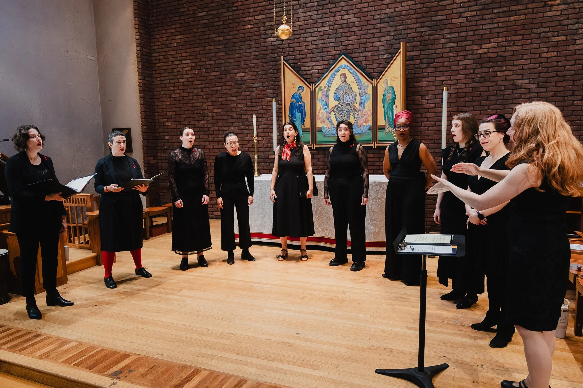 A treble choir in black rehearses in a brick church, spread in a wide arc before an ornate triptych altarpiece, a conductor at a music stand on the right